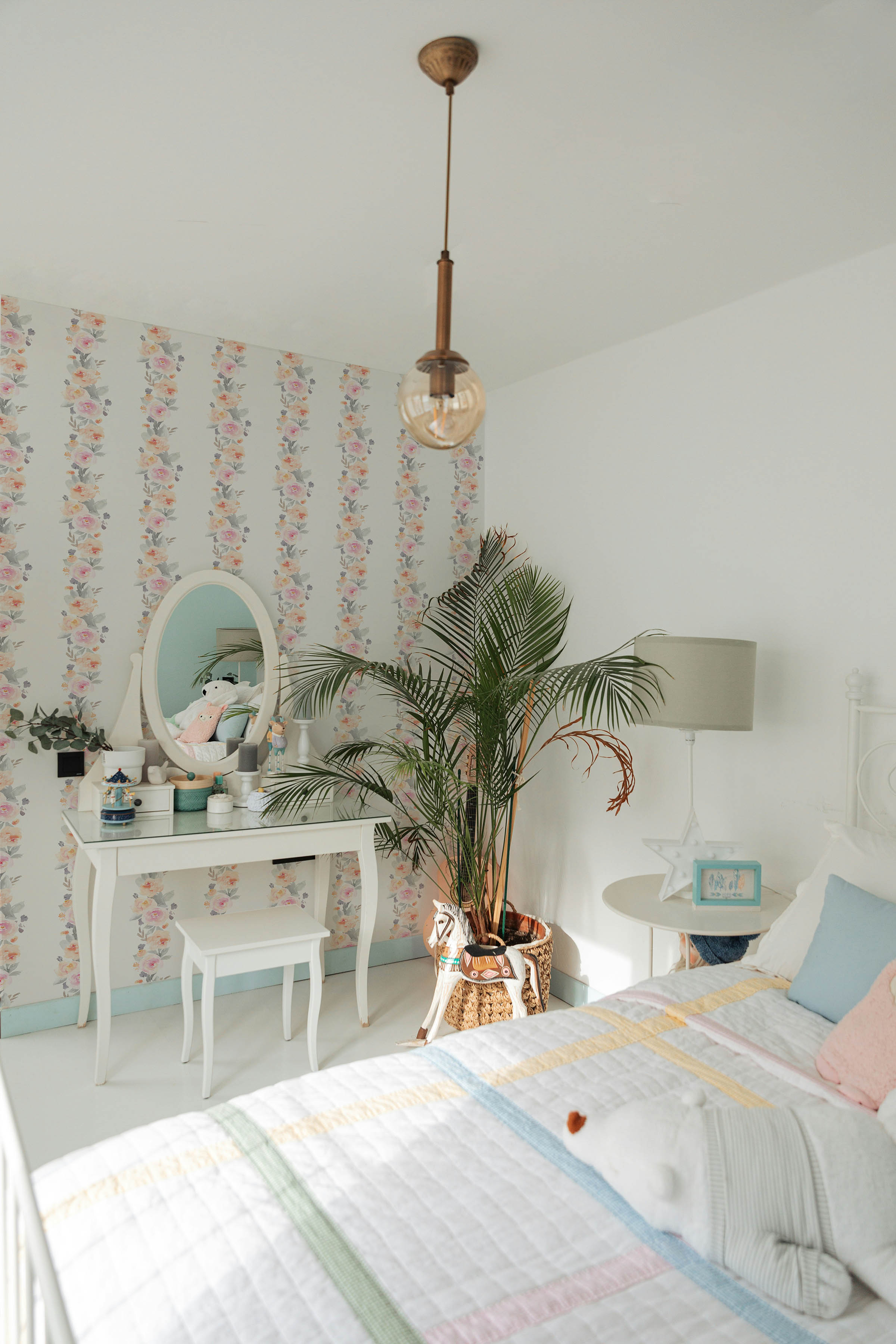 A light and airy bedroom showcases Pastel Dreams Wallpaper adorned with delicate watercolor flowers in shades of pink, peach, and lavender. The room features a bright yellow couch, a white dressing table with a mirror, and a vibrant plant adding a touch of greenery, creating a cheerful and welcoming space.