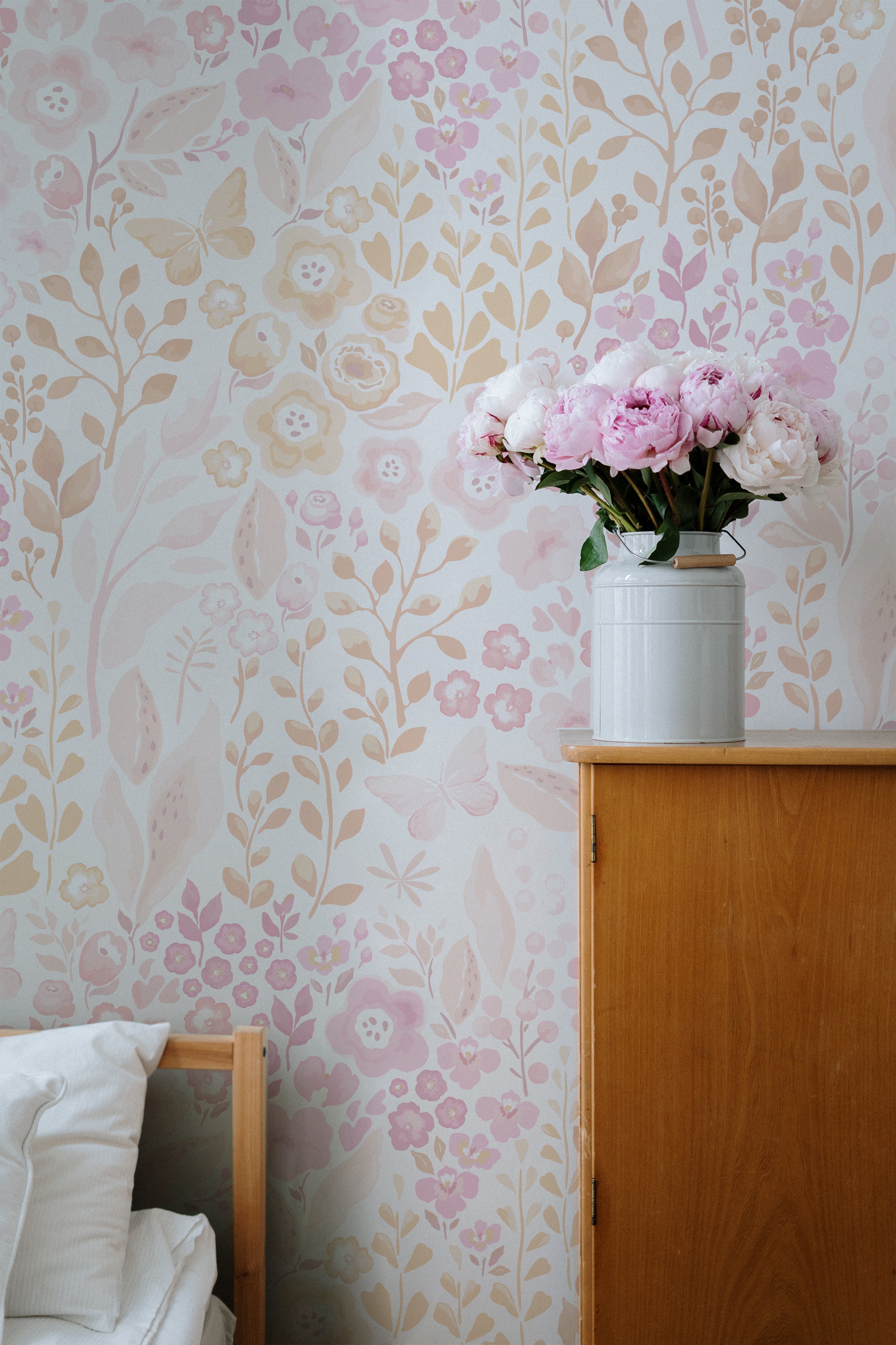 A quaint and peaceful corner of a room featuring the 'Pretty Petals Wallpaper' with soft pink and taupe floral designs. The wallpaper's delicate beauty is complemented by a fresh bouquet of pink peonies in a white vase atop a wooden cabinet, alongside a cushioned chair.