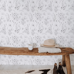 A rustic entryway bench made of a raw wooden plank set against a wall covered in Wildflower Sketch Wallpaper, which features intricate sketches of wildflowers on a white background. On the bench, there's a casual arrangement of a white ceramic mug, a beige felt hat, and a folded knit blanket, giving a sense of welcoming comfort. A pair of well-worn leather boots stands beside the bench on a cowhide rug, completing this homely scene.
