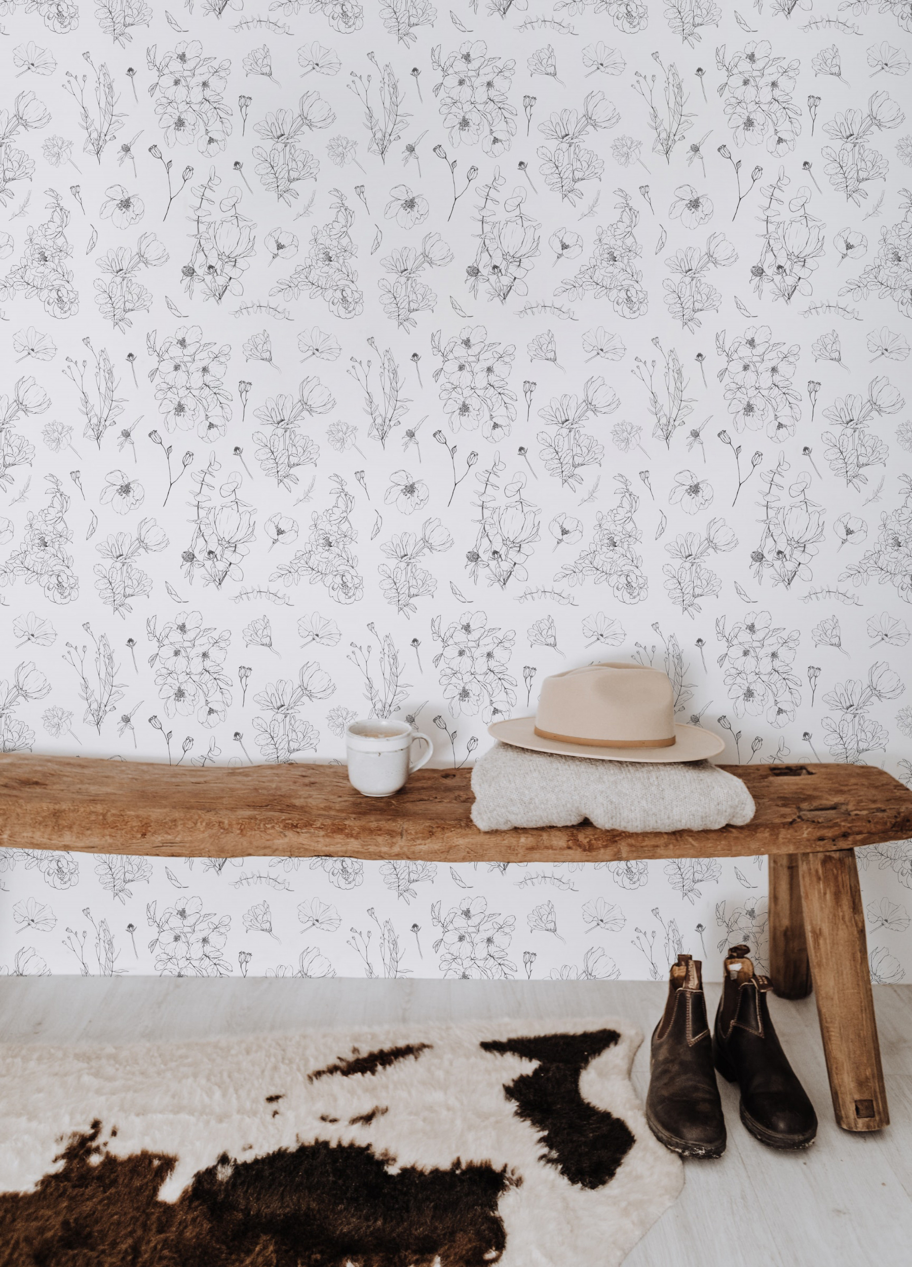 A rustic entryway bench made of a raw wooden plank set against a wall covered in Wildflower Sketch Wallpaper, which features intricate sketches of wildflowers on a white background. On the bench, there's a casual arrangement of a white ceramic mug, a beige felt hat, and a folded knit blanket, giving a sense of welcoming comfort. A pair of well-worn leather boots stands beside the bench on a cowhide rug, completing this homely scene.