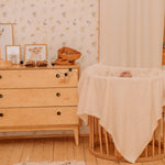 A warmly lit nursery with a soft pink Sunny Floral Wallpaper adorned with delicate blooms and foliage, accompanied by a wooden chest of drawers and a comfy rocking crib with a plush blanket.