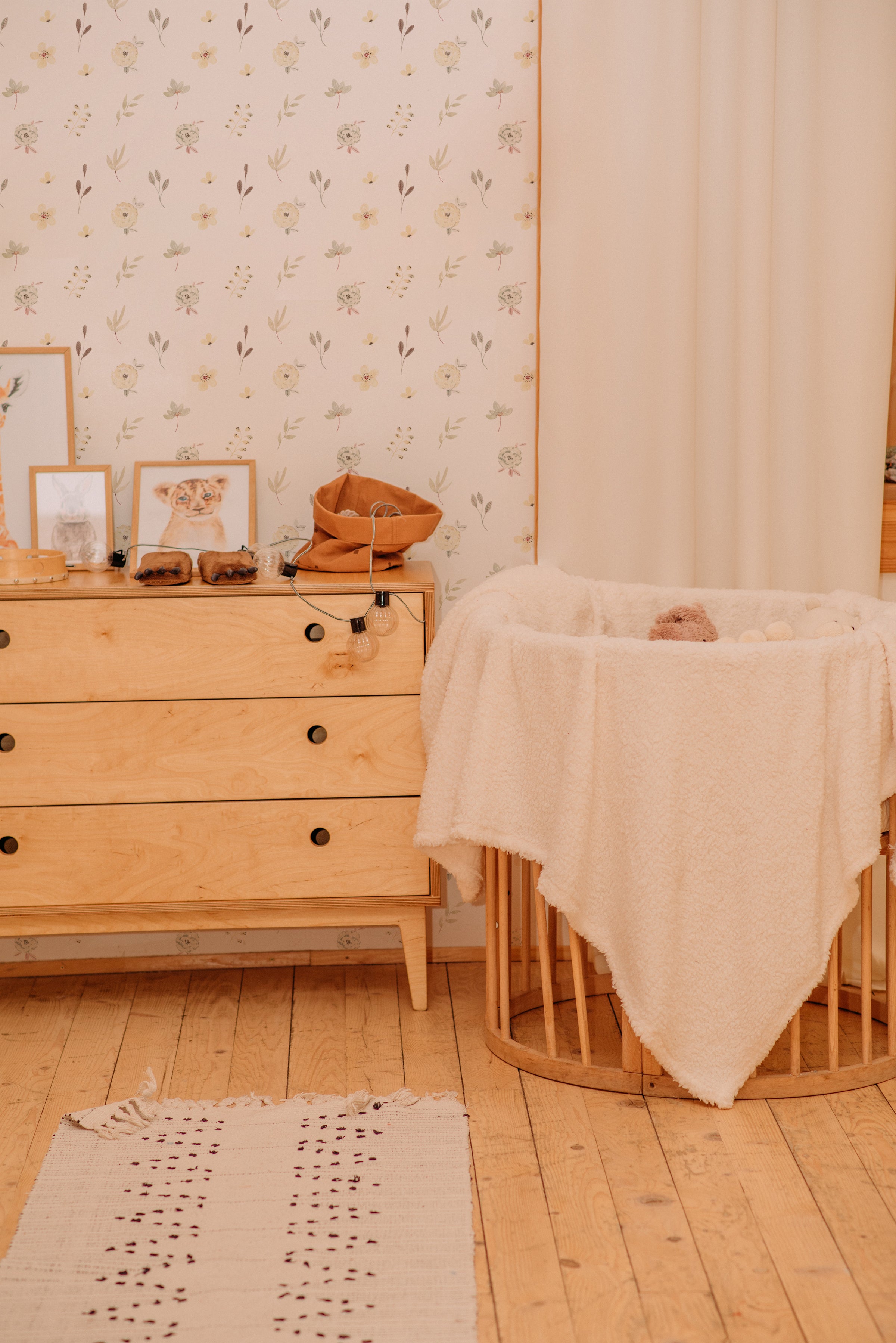 A warmly lit nursery with a soft pink Sunny Floral Wallpaper adorned with delicate blooms and foliage, accompanied by a wooden chest of drawers and a comfy rocking crib with a plush blanket.
