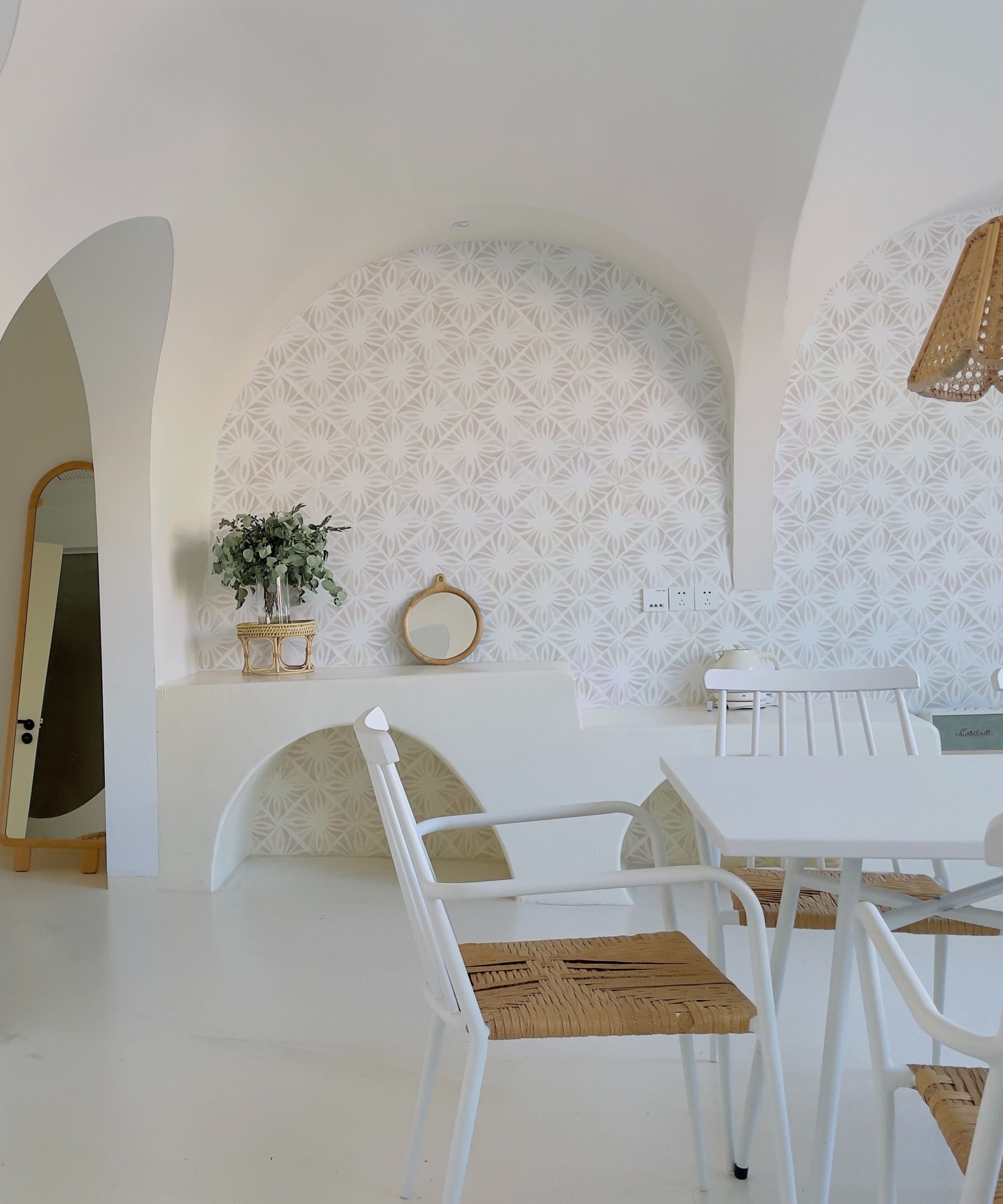 A bright and airy dining nook accented by Moroccan Tile Wallpaper II - Linen, featuring a subtle linen-colored geometric design, paired with modern white chairs and a natural wooden table, all under the charm of a unique arched alcove.