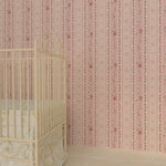 A corner of a room showcasing Vintage Rose Wallpaper with vertical stripes and floral designs in shades of pink and white. A yellow metal crib with a beige and green patterned bedding is partially visible, suggesting a nursery setting.