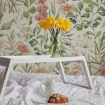 A serene bedroom setting showcasing Watercolour Floral and Leaf Wallpaper II, featuring large, delicate watercolor flowers and leaves on a soft white background. A white bed tray holds a cup and a pastry, complementing the fresh bouquet of yellow daffodils