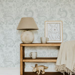 A cozy corner decorated with the Woodland Creatures Wallpaper - Light Sage, adding a touch of whimsy with its delicate drawings of forest fauna. A wooden shelf and a lamp with a natural stone base enhance the wallpaper's rustic and tranquil vibe