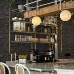 A stylish café interior featuring Black Brick Wallpaper with a textured, stacked stone design in dark gray. The wallpaper adds depth and sophistication to the space, highlighted by brass shelving, white counter stools, and a marble countertop with various coffee-making equipment and decor items.