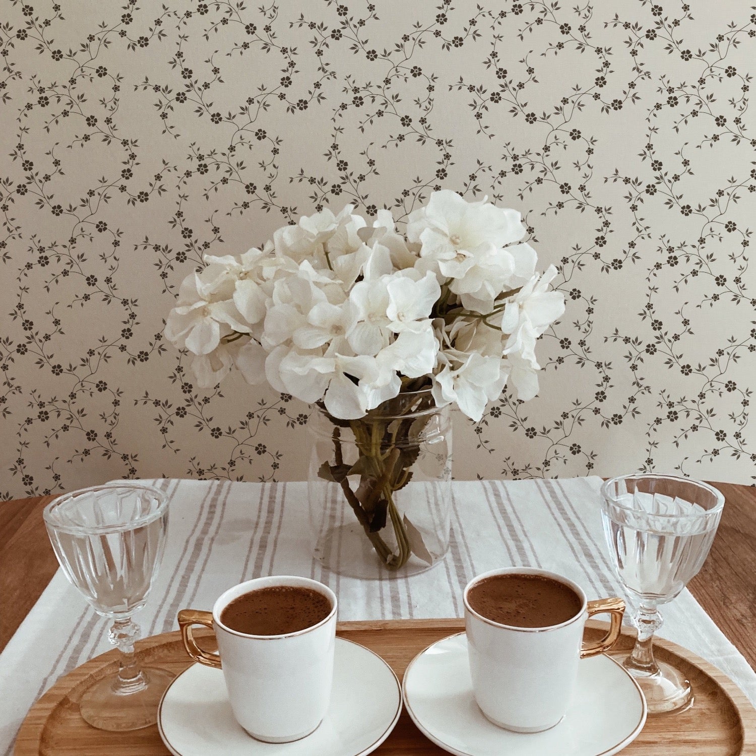 An inviting dining scene featuring the "Mini Charming Floral Wallpaper - Deep Brown" on the wall. The charming floral pattern provides a quaint backdrop for a simple wooden dining table with a glass vase of white hydrangeas, two cups of coffee, and elegant glass stemware, highlighting the wallpaper's ability to enhance a homely dining experience.