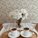 An inviting dining scene featuring the "Mini Charming Floral Wallpaper - Deep Brown" on the wall. The charming floral pattern provides a quaint backdrop for a simple wooden dining table with a glass vase of white hydrangeas, two cups of coffee, and elegant glass stemware, highlighting the wallpaper's ability to enhance a homely dining experience.
