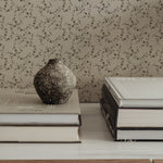 A close-up image emphasizing the fine details of the "Mini Charming Floral Wallpaper - Deep Brown". The pattern consists of tiny brown floral designs with fine green stems and leaves on a light background. The wallpaper serves as a backdrop to a shelf with neatly stacked books, topped by a textured ceramic vase, exemplifying the wallpaper's suitability for a refined and literary space.