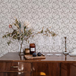 A cozy interior setting showcasing the "Mini Charming Floral Wallpaper - Deep Brown" as a backdrop. The wallpaper has a delicate pattern of small brown flowers with subtle green leaves on an off-white background. The scene includes a wooden sideboard with a clear vase holding dried flowers, figs on a wooden tray, and glassware, creating a warm and inviting ambiance.