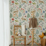 A whimsical children's room corner with Colourful Spring Bunnies Wallpaper featuring playful rabbits and a mix of floral and botanical prints. A wooden chair with a woven seat holds a stuffed rabbit, set against a backdrop of light beige wallpaper adorned with pastel-colored flowers and green foliage.