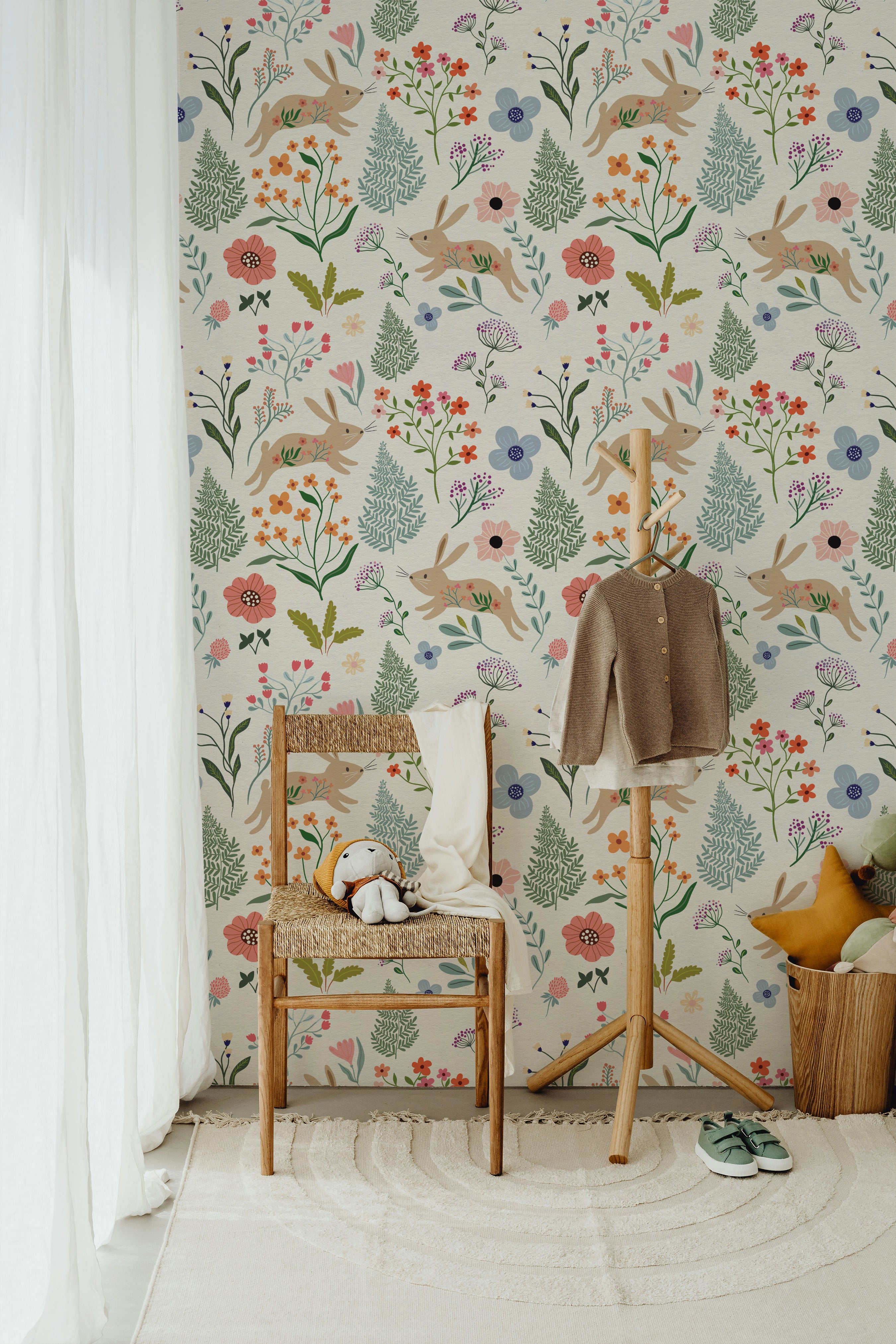 A whimsical children's room corner with Colourful Spring Bunnies Wallpaper featuring playful rabbits and a mix of floral and botanical prints. A wooden chair with a woven seat holds a stuffed rabbit, set against a backdrop of light beige wallpaper adorned with pastel-colored flowers and green foliage.