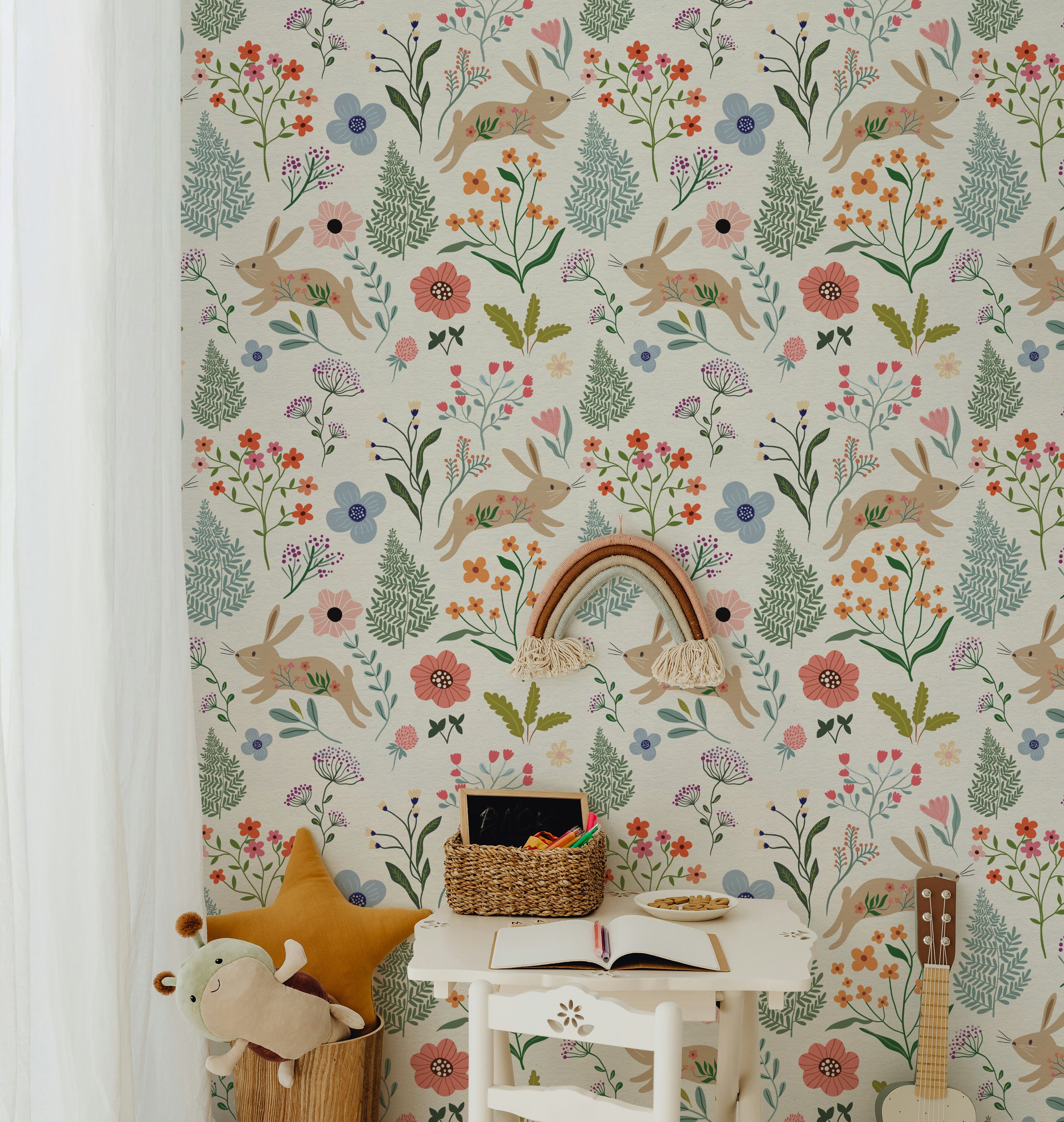 A cozy reading nook for children decorated with Colourful Spring Bunnies Wallpaper. The wallpaper's delightful pattern of rabbits and varied flowers creates a lively and engaging environment. A small white table holds books and toys, enhancing the playful theme.