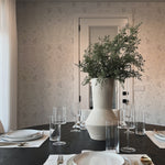 A beautifully set dining table in a room adorned with Rustic Vintage Floral wallpaper, featuring subtle floral patterns that provide a timeless charm, complemented by a large vase of greenery on the table, enhancing the room's rustic elegance.