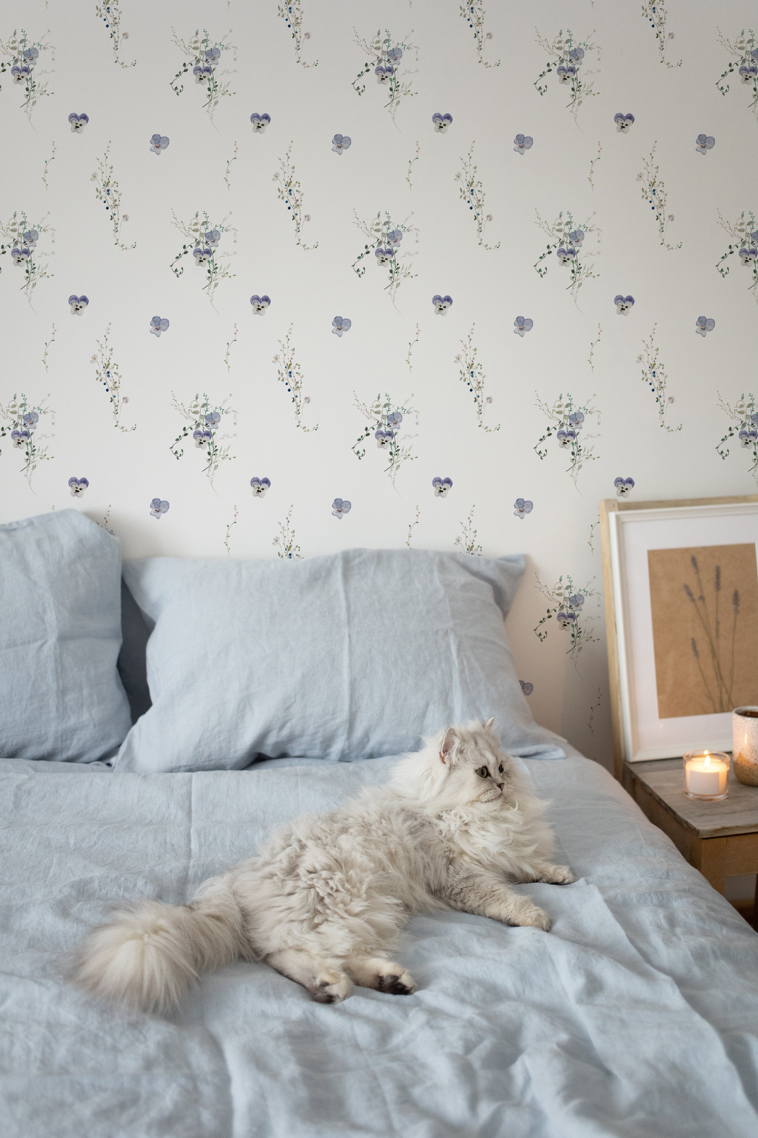A bedroom with soft blue bedding matching the Delicate Blue Wildflower Wallpaper on the wall. The wallpaper features sprigs of blue wildflowers with green leaves, adding a serene and natural touch. A fluffy white cat lounges on the bed, blending in with the peaceful decor.