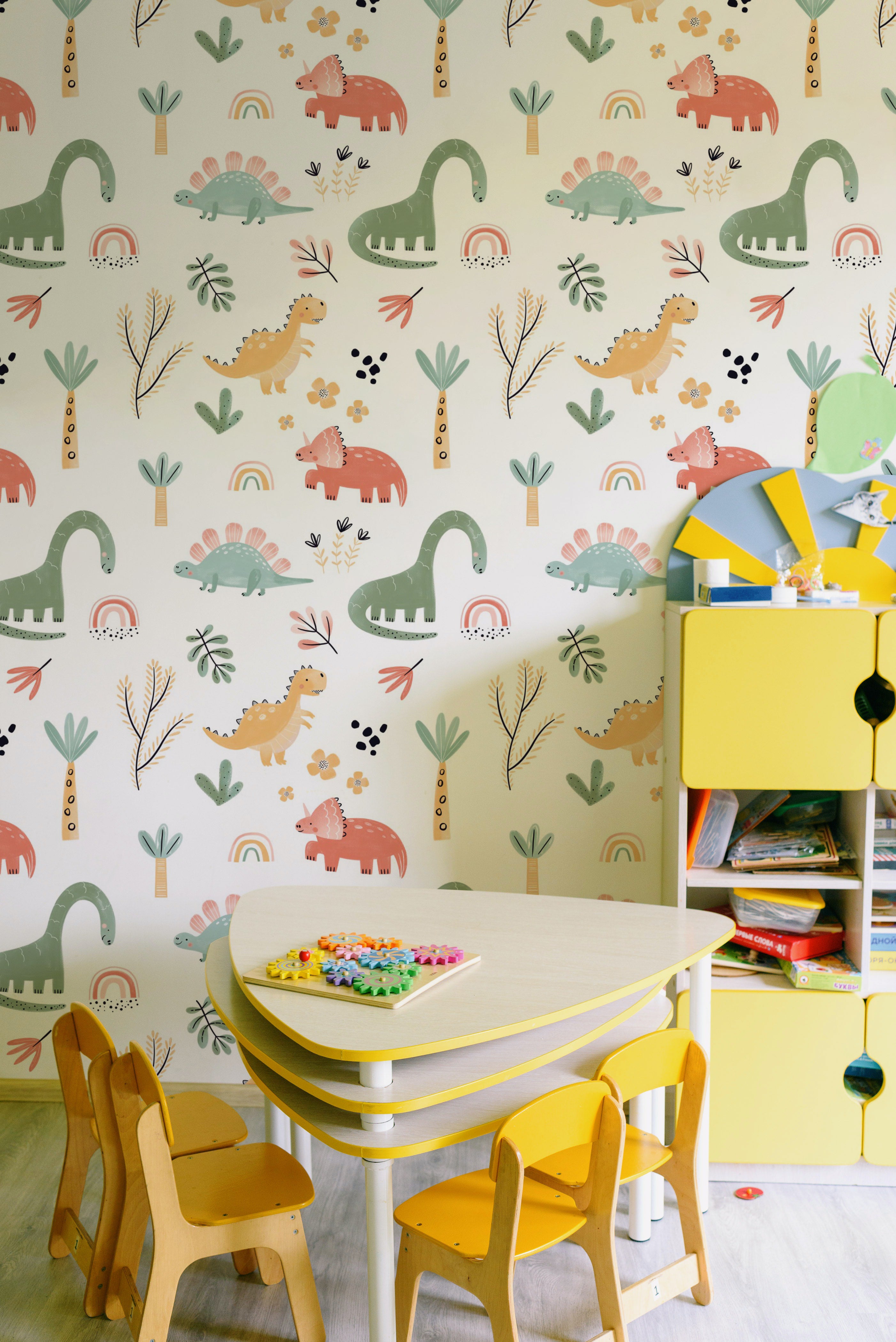 A vibrant children's room corner with Dinosaur Nursery Wallpaper enveloping the space in a scene of prehistoric playfulness, complemented by bright yellow storage and toys, which add a lively contrast to the gentle wall pattern.