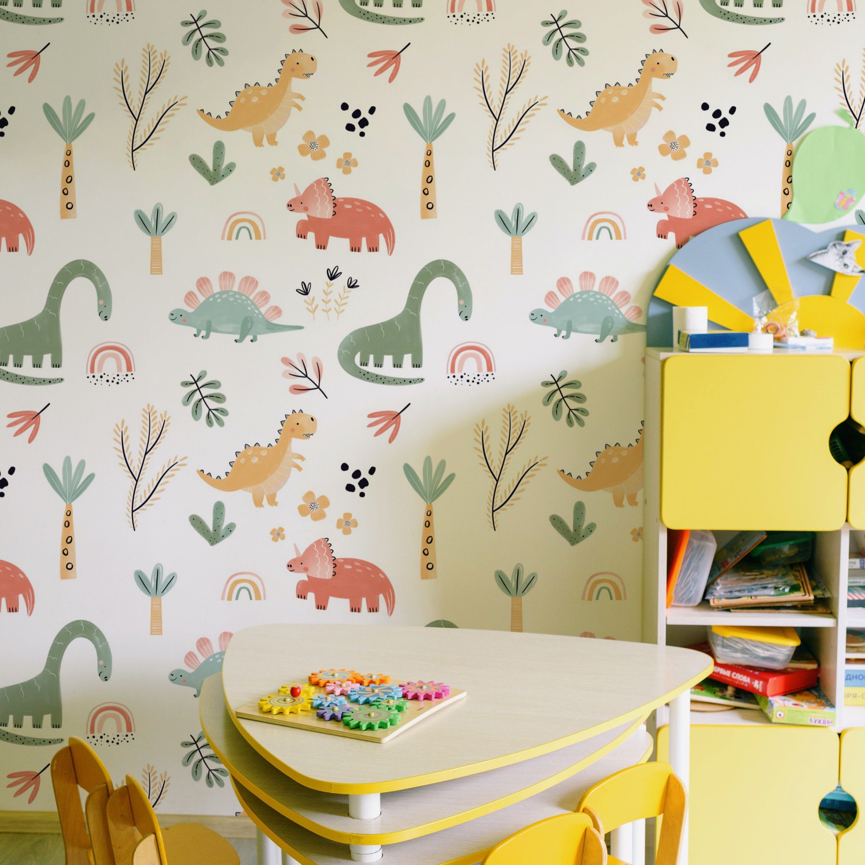 A playful and colorful children's play area featuring Dinosaur Nursery Wallpaper, with charming illustrations of friendly dinosaurs and flora in pastel colors, paired with a yellow and white children's table and chairs, creating a whimsical space for creativity and play.