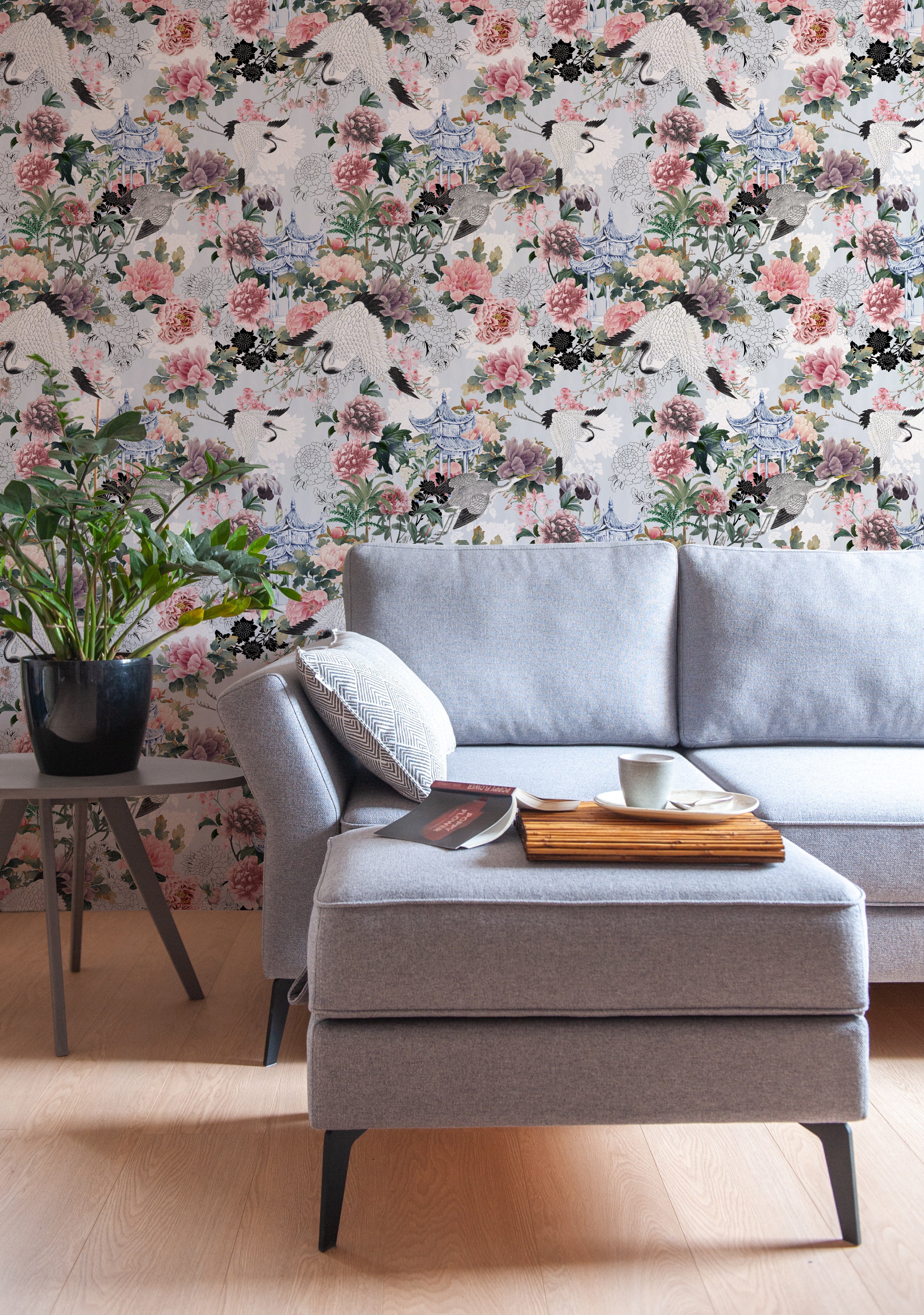 A stylish living room enhanced by the Kyoto Dreams Wallpaper, creating an elegant focal wall behind a contemporary grey sofa. The wallpaper's design, depicting large peonies and cranes amidst traditional Japanese architecture, infuses the space with a sense of luxury and cultural beauty
