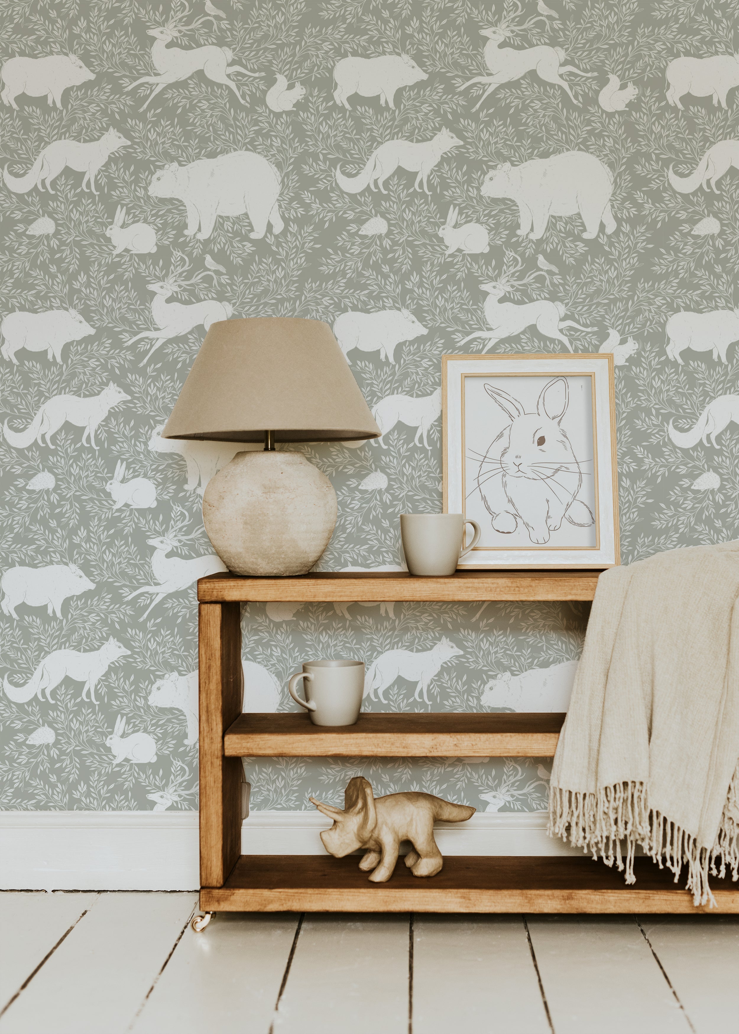 A cozy living room setting with a wooden shelf, ceramic lamp, and framed rabbit illustration in front of a green wallpaper featuring white woodland animals such as bears, foxes, and rabbits