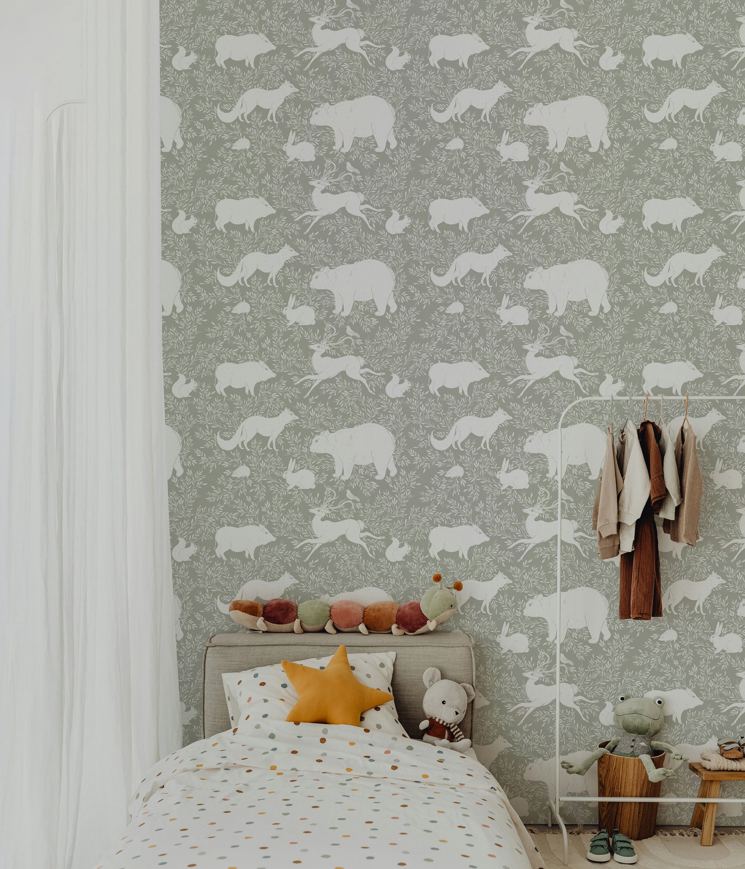 A children's bedroom with a bed dressed in polka dot bedding and stuffed animals, featuring a green wallpaper with white woodland animals like bears, foxes, and rabbits, creating a whimsical and calming atmosphere.