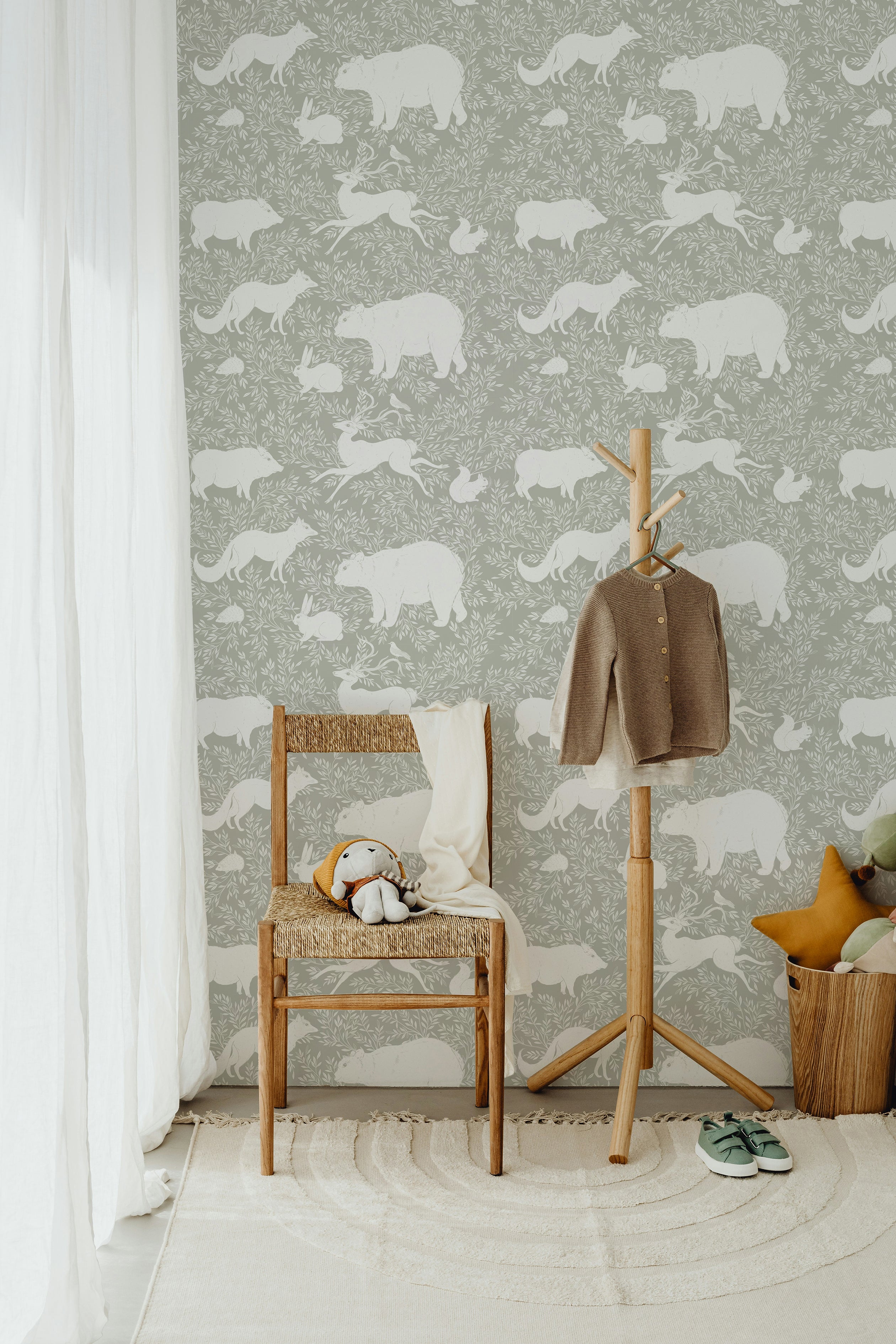 A serene corner with a wooden chair, soft toys, and a hanging coat against a green wallpaper decorated with white woodland animals such as bears, foxes, and rabbits, evoking a peaceful woodland retreat.