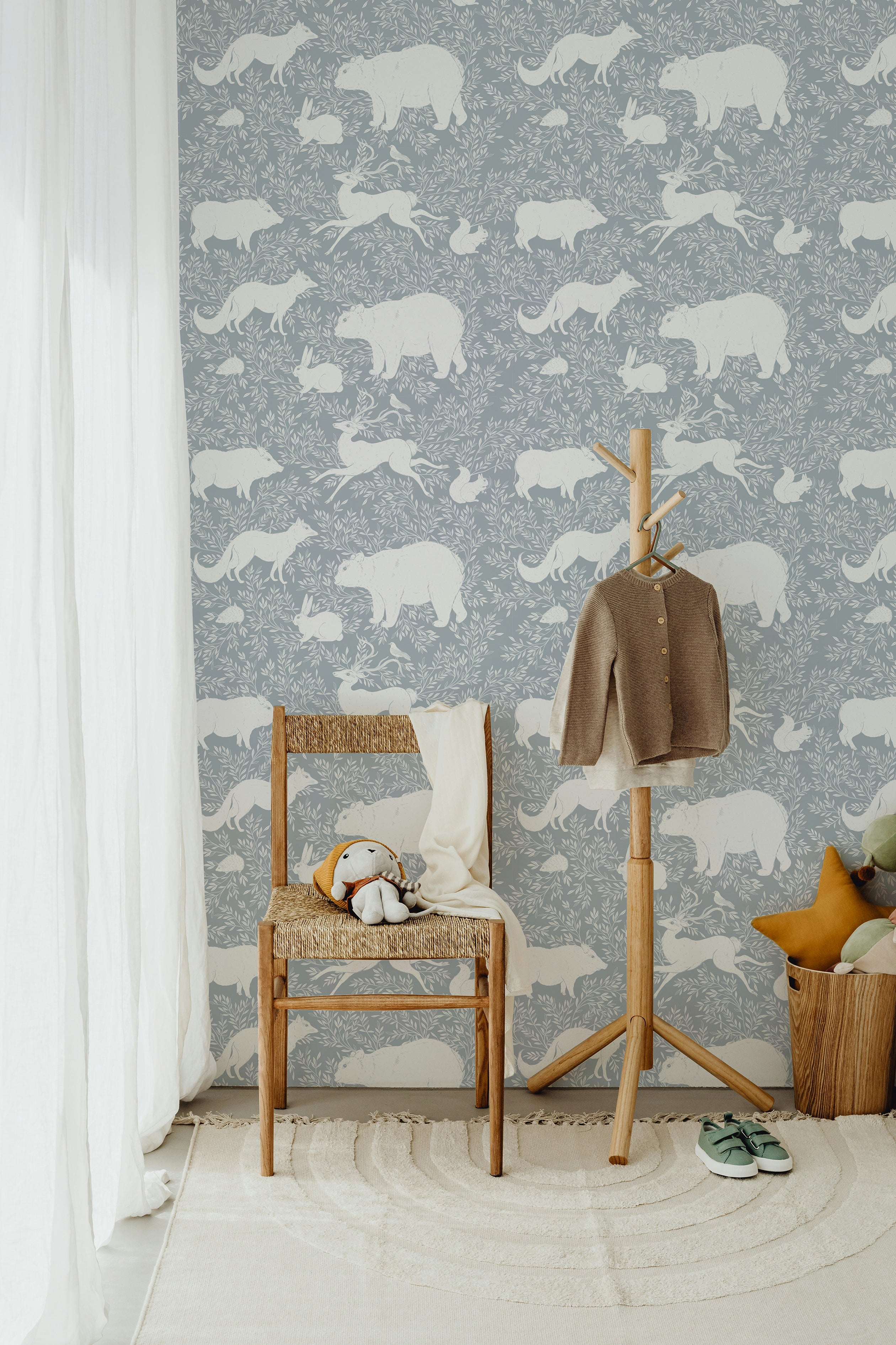 A serene corner with a wooden chair, soft toys, and a hanging coat against a pale blue wallpaper decorated with white woodland animals such as bears, foxes, and rabbits, evoking a peaceful woodland retreat.