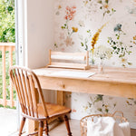 A cozy workspace set against a backdrop of Fiori Wallpaper, featuring gentle floral motifs in watercolor. The natural light from the window illuminates the subtle colors of the wallpaper, paired with a simple wooden desk and a traditional wooden chair