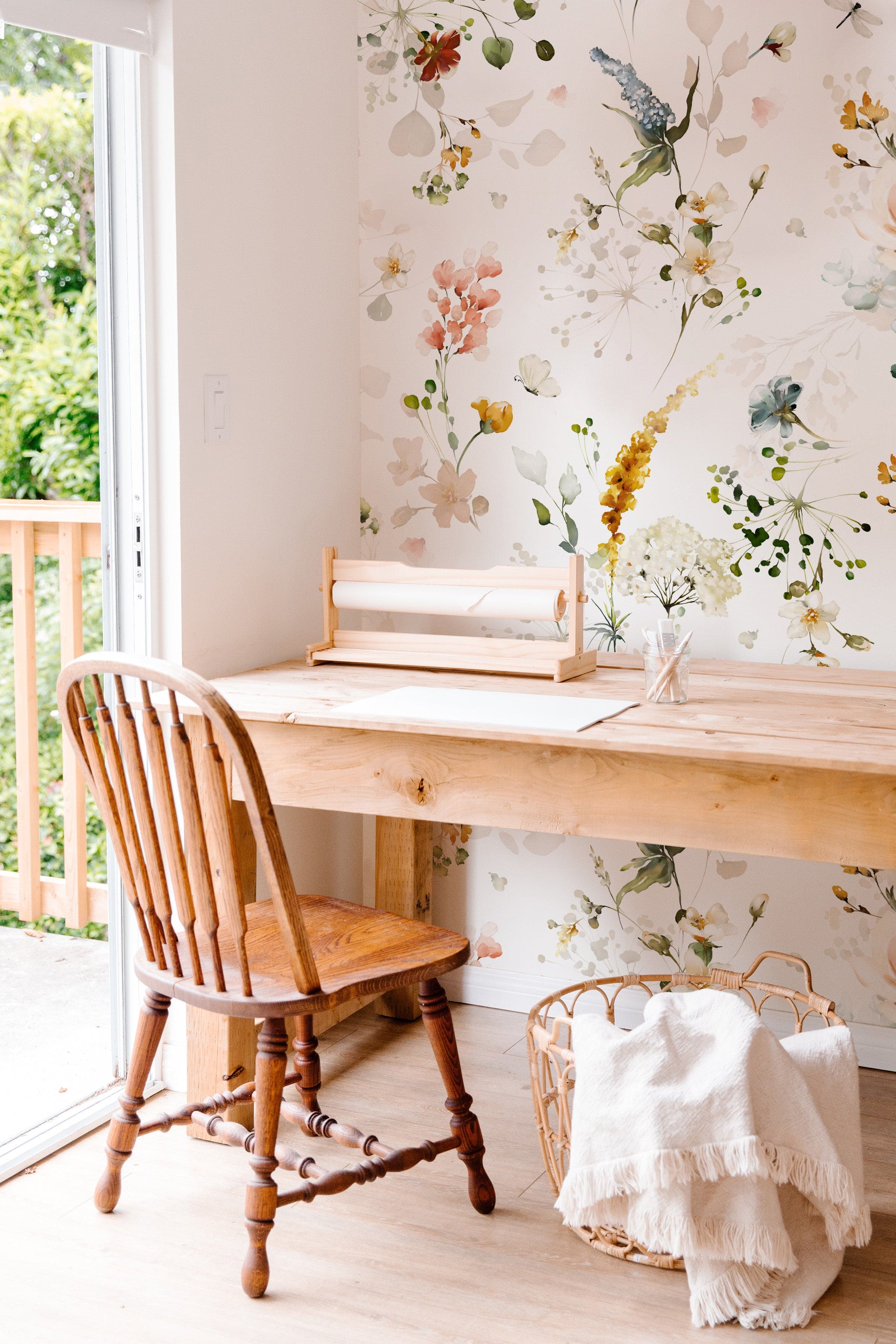 A cozy workspace set against a backdrop of Fiori Wallpaper, featuring gentle floral motifs in watercolor. The natural light from the window illuminates the subtle colors of the wallpaper, paired with a simple wooden desk and a traditional wooden chair