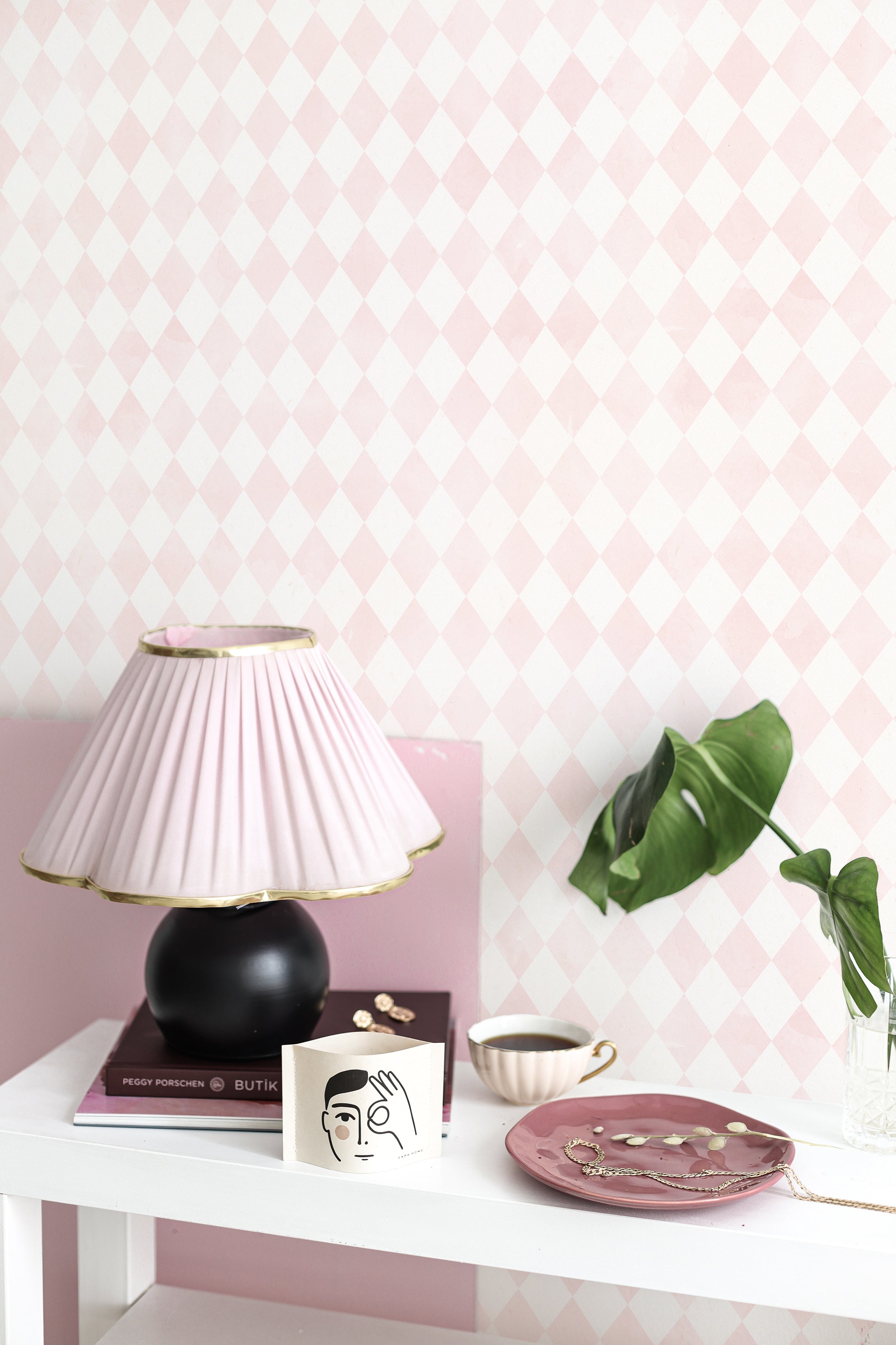 A stylish corner of a room showcasing Rustic Watercolour Geometric Diamonds Wallpaper. The light pink and white diamond pattern provides a soft, watercolor-like background for a black table lamp, books, and decorative items, enhancing the chic, feminine decor.
