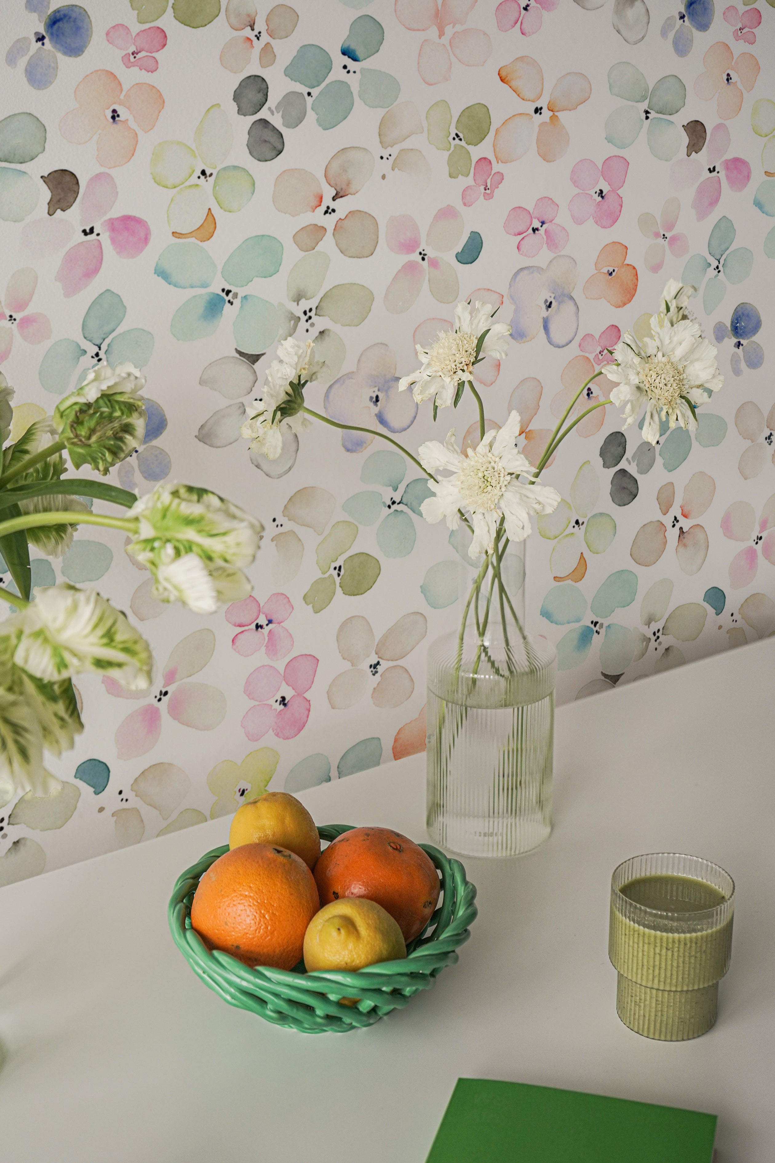 A modern workspace is given a splash of color with the Hand Painted Floral Wallpaper, featuring a myriad of watercolor flowers in pastel shades. A white desk with fresh flowers, a bowl of citrus fruit, and a light green book create a scene of vibrant yet serene productivity.