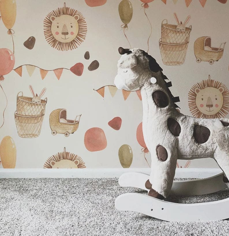 A cozy corner of a nursery showing a part of the wall decorated with a cute, animal-themed wallpaper. Visible motifs include lions' faces, balloons, and bunting flags, complemented by a soft plush rocking giraffe in the foreground, enhancing the playful, child-friendly decor.