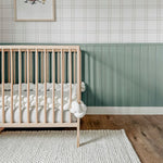 Detailed view of a nursery corner with Traditional Tartan Plaid Wallpaper and green wainscoting. The room is furnished with a crib filled with plush toys, under a framed playful artwork, enhancing the room's warm and welcoming vibe.