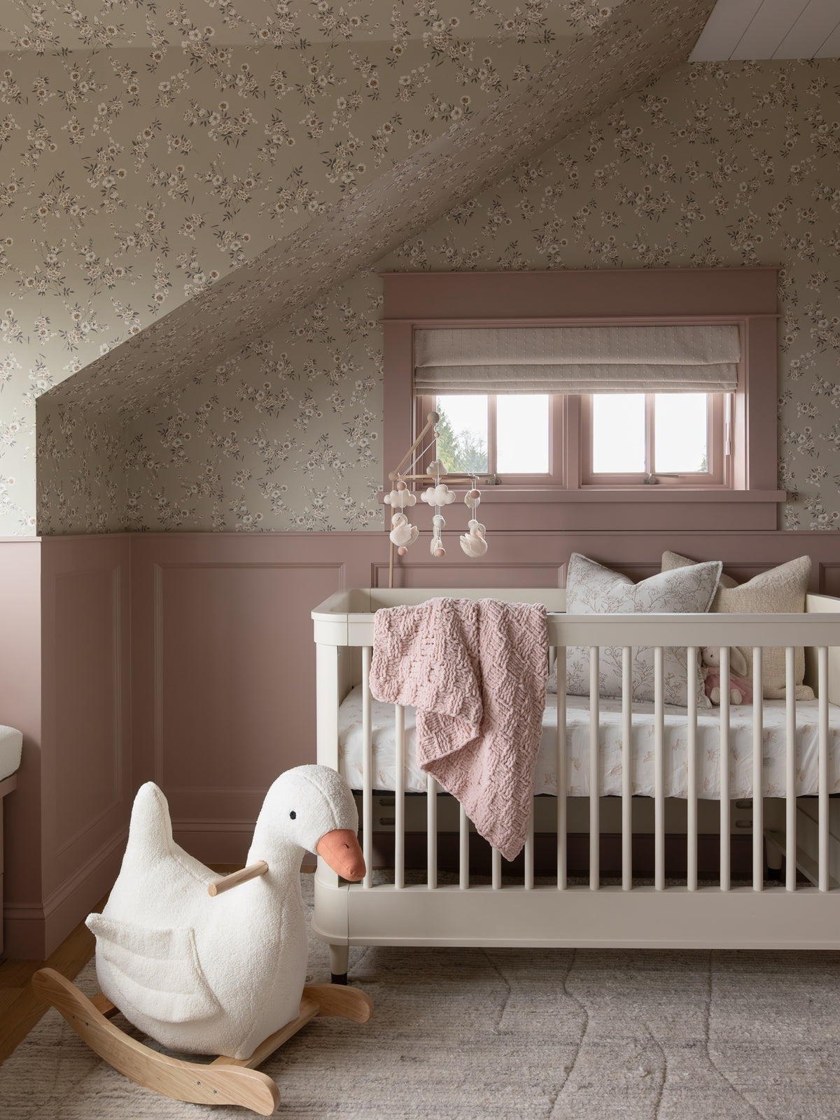 A styled nursery room featuring a white wooden crib with a dusty pink blanket, a white rocking duck toy, and floral wallpaper on the walls.
