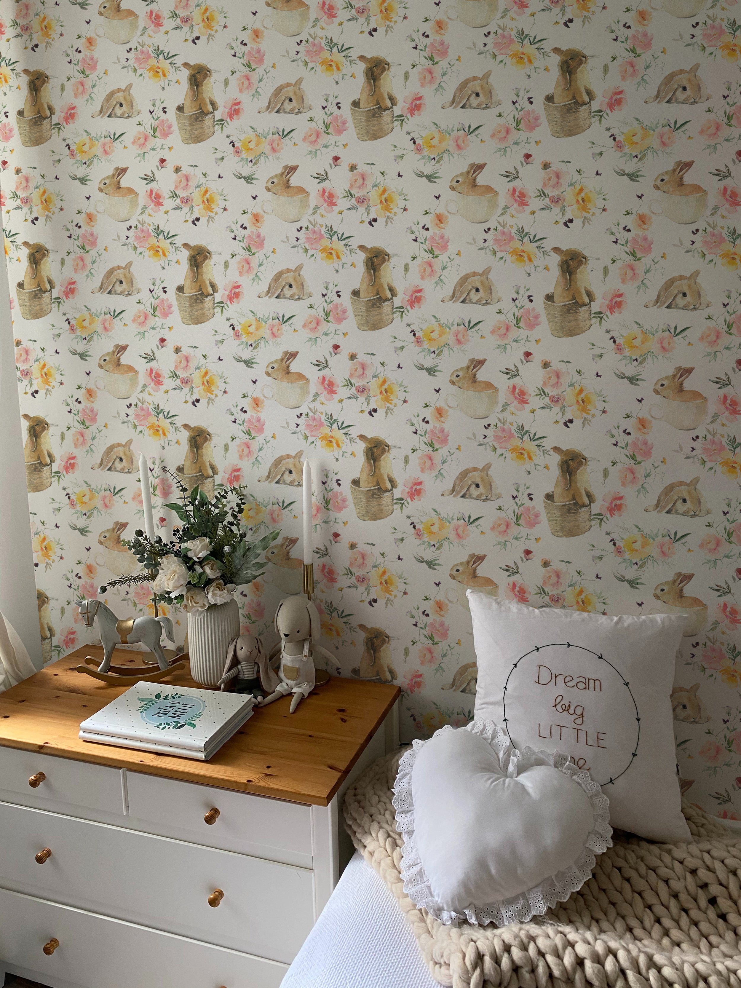 A charming child's room adorned with Garden Bunnies Wallpaper showcasing adorable rabbits and colorful flowers. The decor includes a white dresser topped with plush toys and children's books, enhancing the room's whimsical feel.