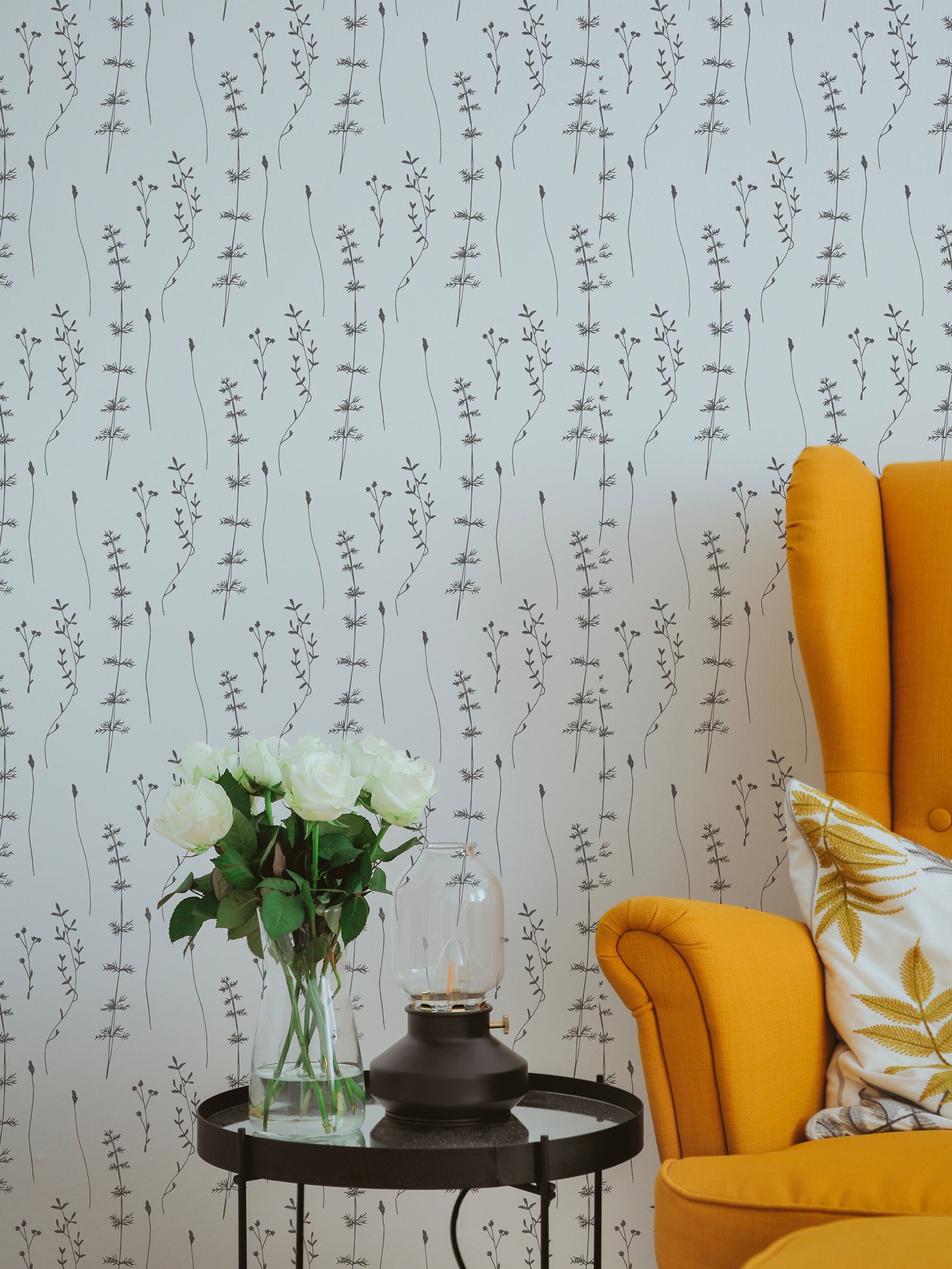 A cozy corner in a living room adorned with Delicate Wildflower Wallpaper featuring subtle, hand-drawn wildflower motifs in black on a soft gray background. A yellow upholstered chair and a round black side table with a vase of white roses and a modern lantern add to the charm of the space.