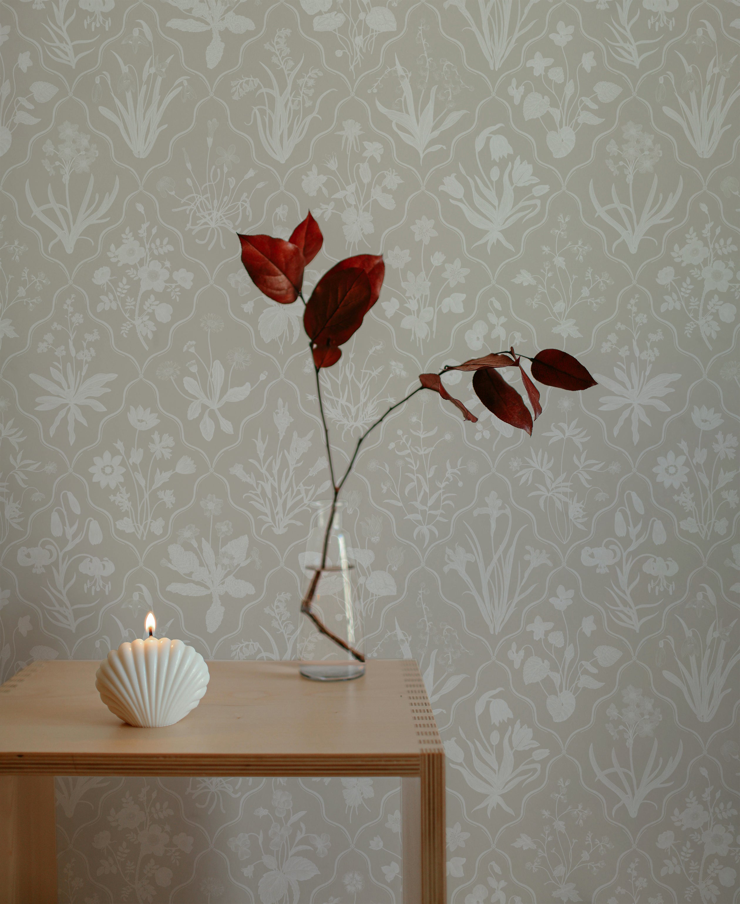 Mille-Fleurs Wallpaper II in a light beige shade with a floral pattern, displayed with minimalist decor including a candle and a branch in a vase, showcasing its elegant design