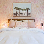 A charming bedroom adorned with 'Pretty Petals Wallpaper' creating a soft, romantic backdrop behind a bed dressed in white linens. The decor is completed with pink throw pillows, elegant bedside lamps, and a framed photograph of palm trees.