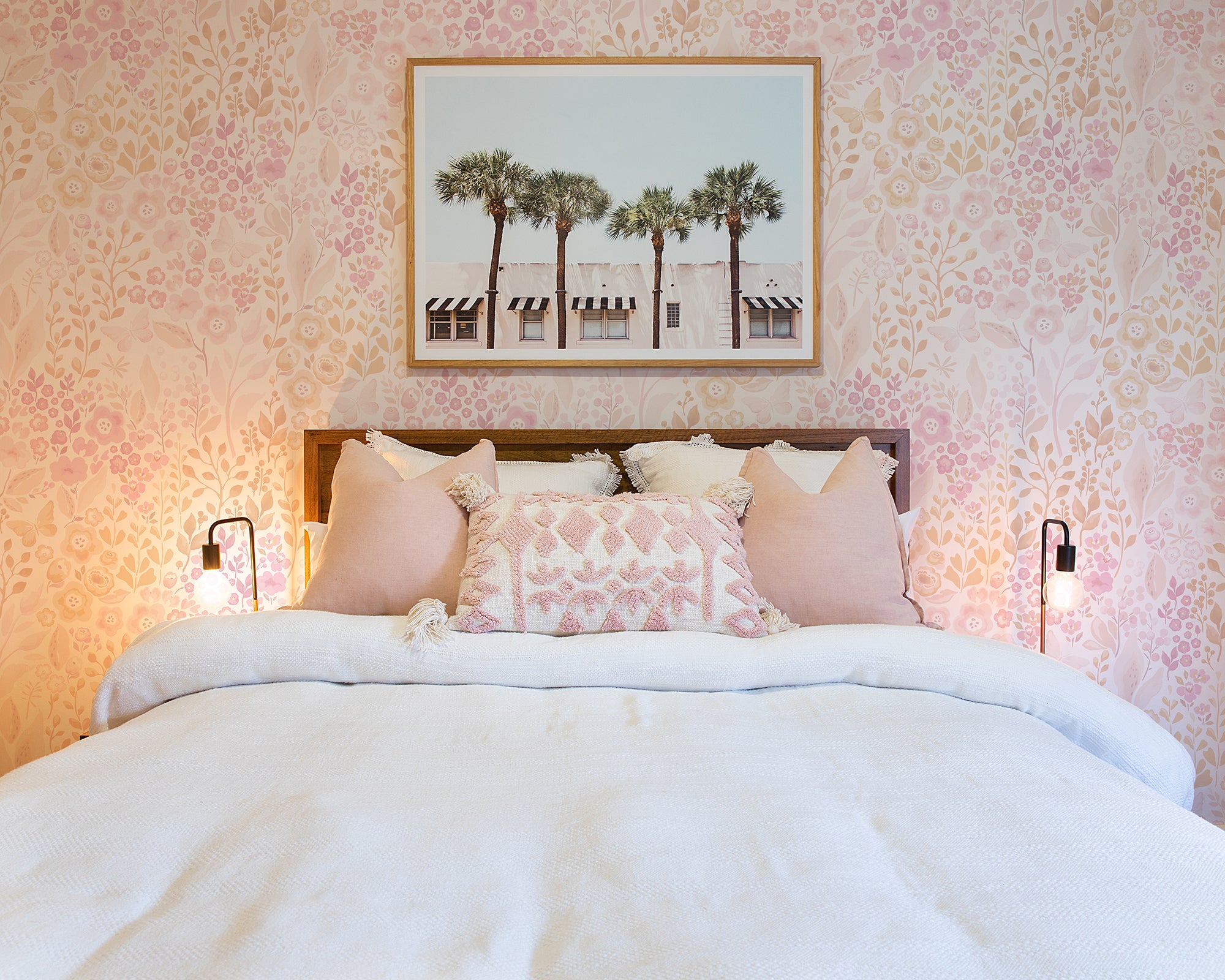 A charming bedroom adorned with 'Pretty Petals Wallpaper' creating a soft, romantic backdrop behind a bed dressed in white linens. The decor is completed with pink throw pillows, elegant bedside lamps, and a framed photograph of palm trees.
