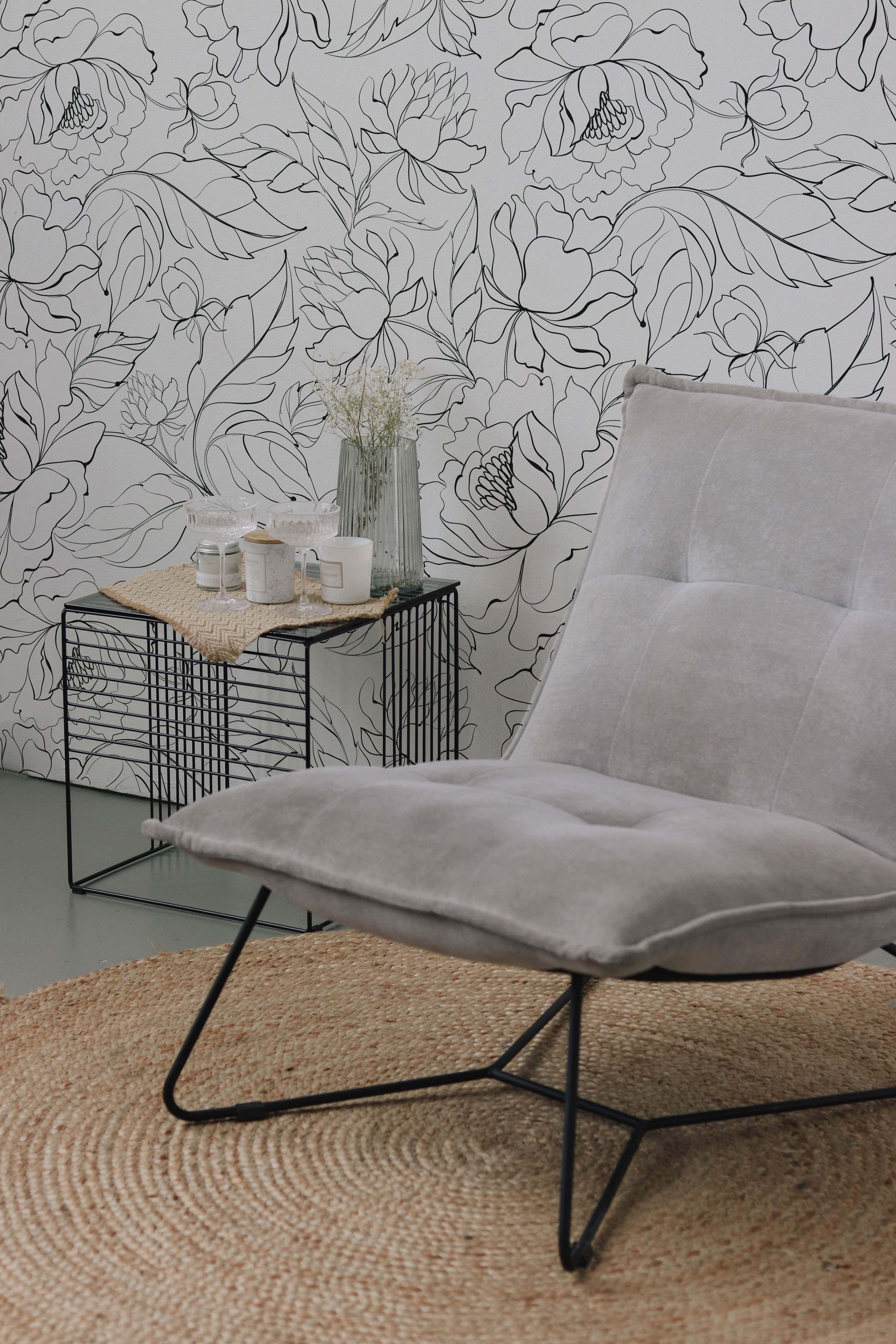 A close-up view of a stylish interior corner, with modern black floral wallpaper featuring a detailed floral line art, accompanied by a trendy wire table holding candles and a glass vase, set against a textured gray chair.
