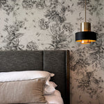 A modern bedroom pairs the Toile De Jouy Monochrome Floral Wallpaper with contemporary furnishings, including a dark upholstered bed and a chic pendant light, blending traditional and current styles