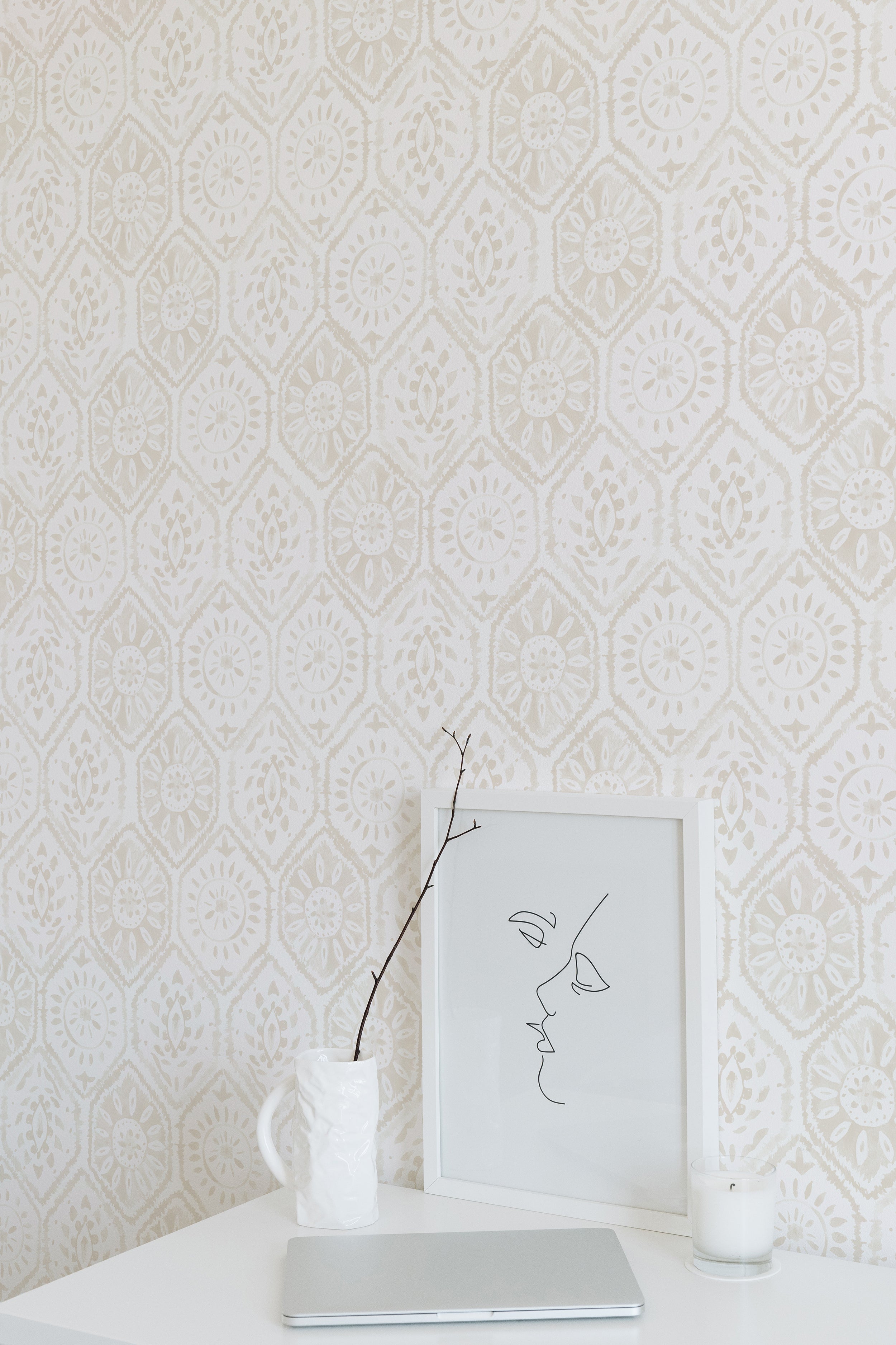 A minimalist workspace highlighted by the Moroccan Tile Wallpaper in Ecru. The wallpaper's intricate pattern provides a soft, decorative background that pairs well with the simple desk, modern line art in a frame, a white laptop, and a single branch in a vase, creating a serene and stylish area.