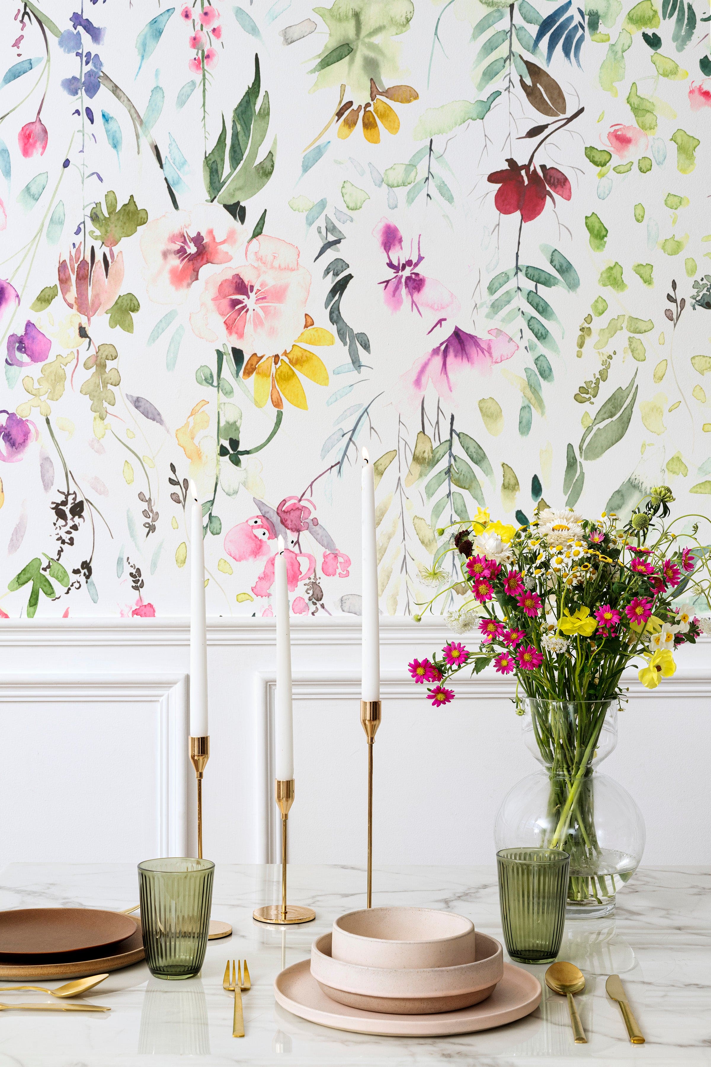 A sophisticated dining area with Hera's Floral Wallpaper as a backdrop. The wall is adorned with this vivid floral pattern, setting a festive and elegant atmosphere for the dining space, complete with tall white candles and a bouquet of fresh flowers.