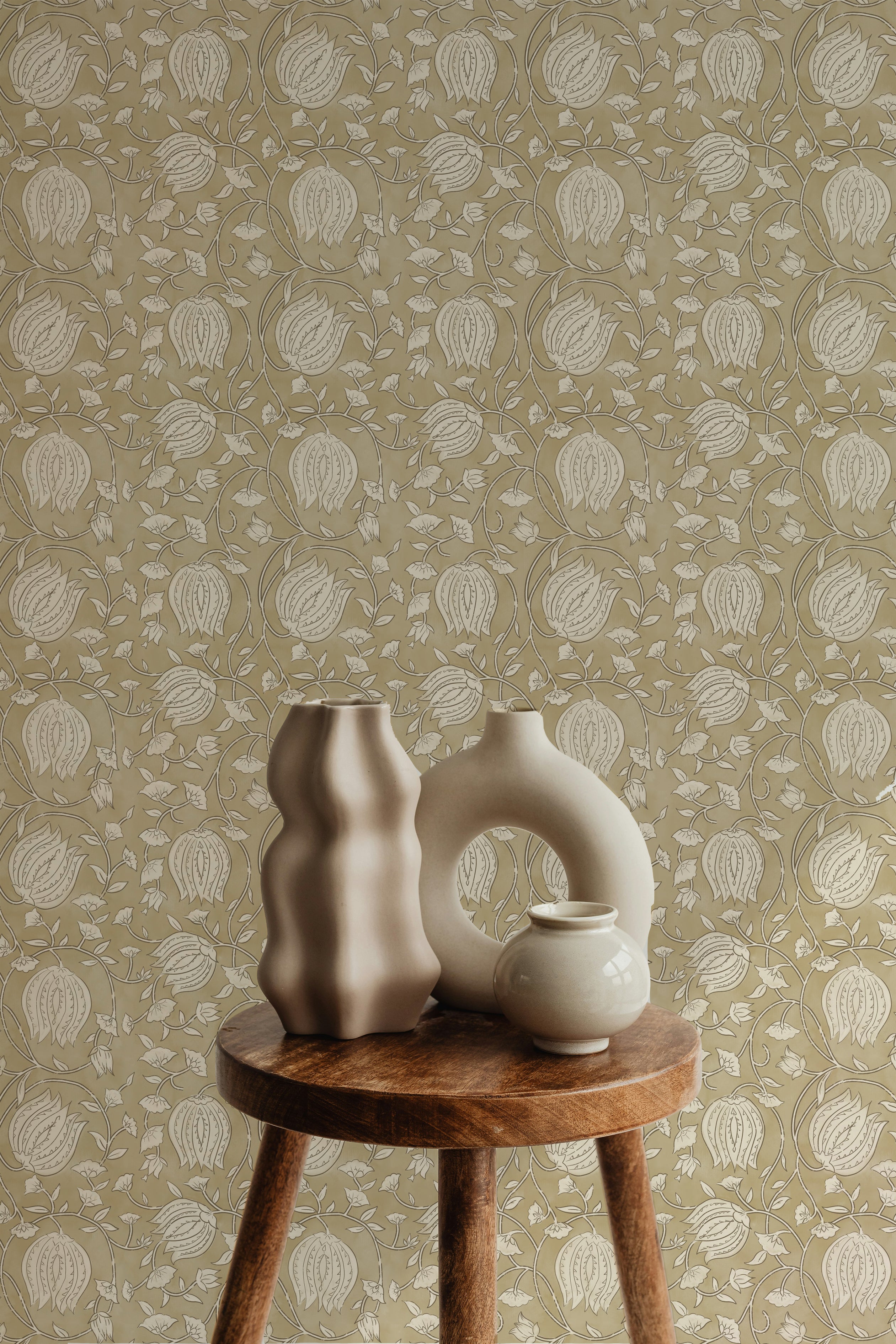Artistic display against a mustard-colored Cornflower Wallpaper, showcasing an intricate design of beige and off-white floral motifs. The composition includes two unique ceramic vases on a rustic wooden stool, enhancing the vintage aesthetic of the space.