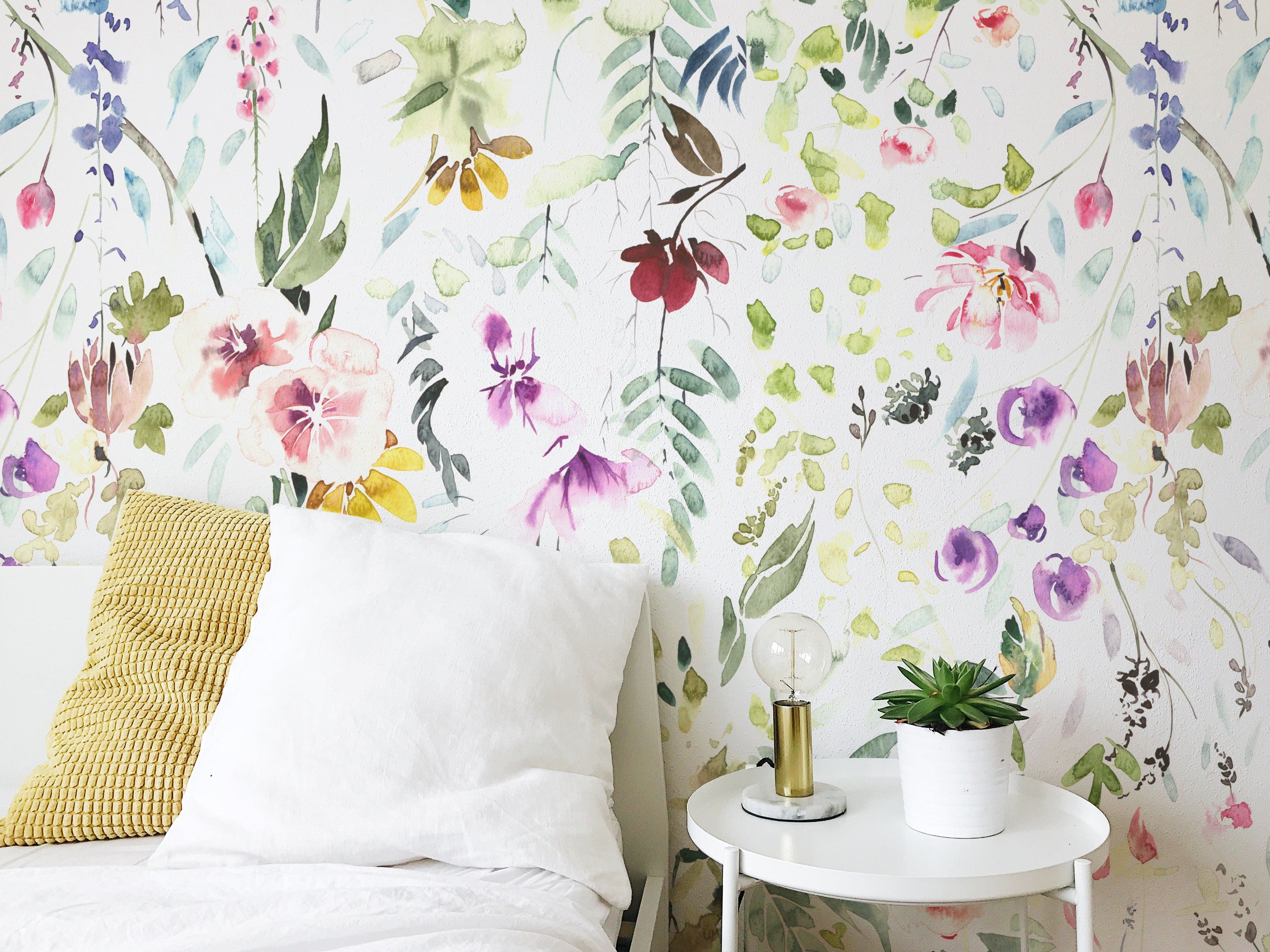 A charming bedroom setting showcasing Hera's Floral Wallpaper, with a vibrant and colorful watercolor floral pattern. The wallpaper provides a lively backdrop to a simple white bed with beige pillows, complemented by a small white side table with a green potted plant and a unique lamp.