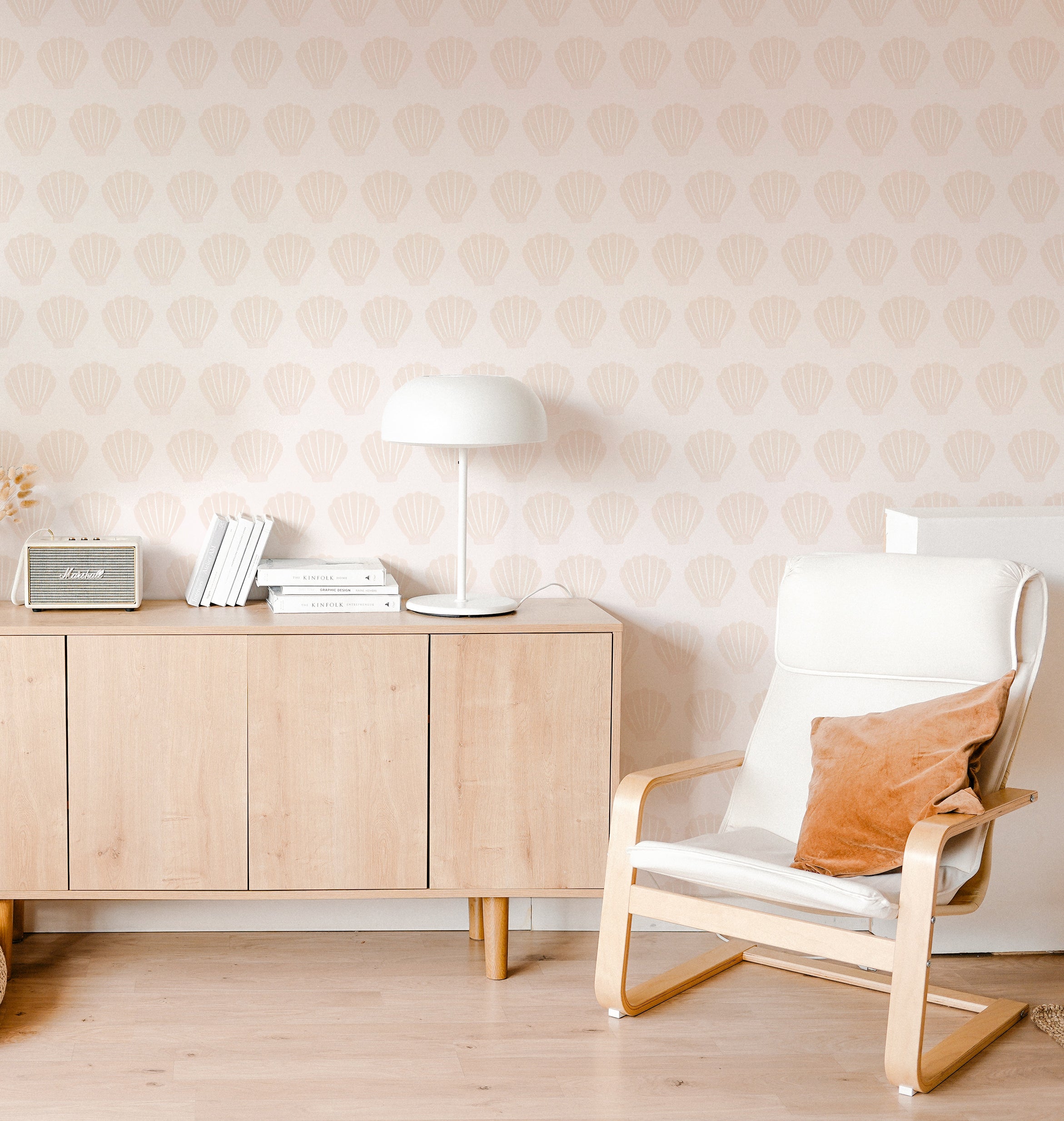 A cozy living room featuring a Scandinavian style decor with Mermaid Sea Shell wallpaper in soft pink, displaying elegant shell patterns. A modern white chair and a wooden cabinet with books and a vintage radio enhance the serene atmosphere.