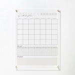 The full acrylic calendar board is shown mounted on a white wall with gold pins at each corner. The board features the same monthly grid and sections for weekly notes and reminders, maintaining its elegant and clean design.