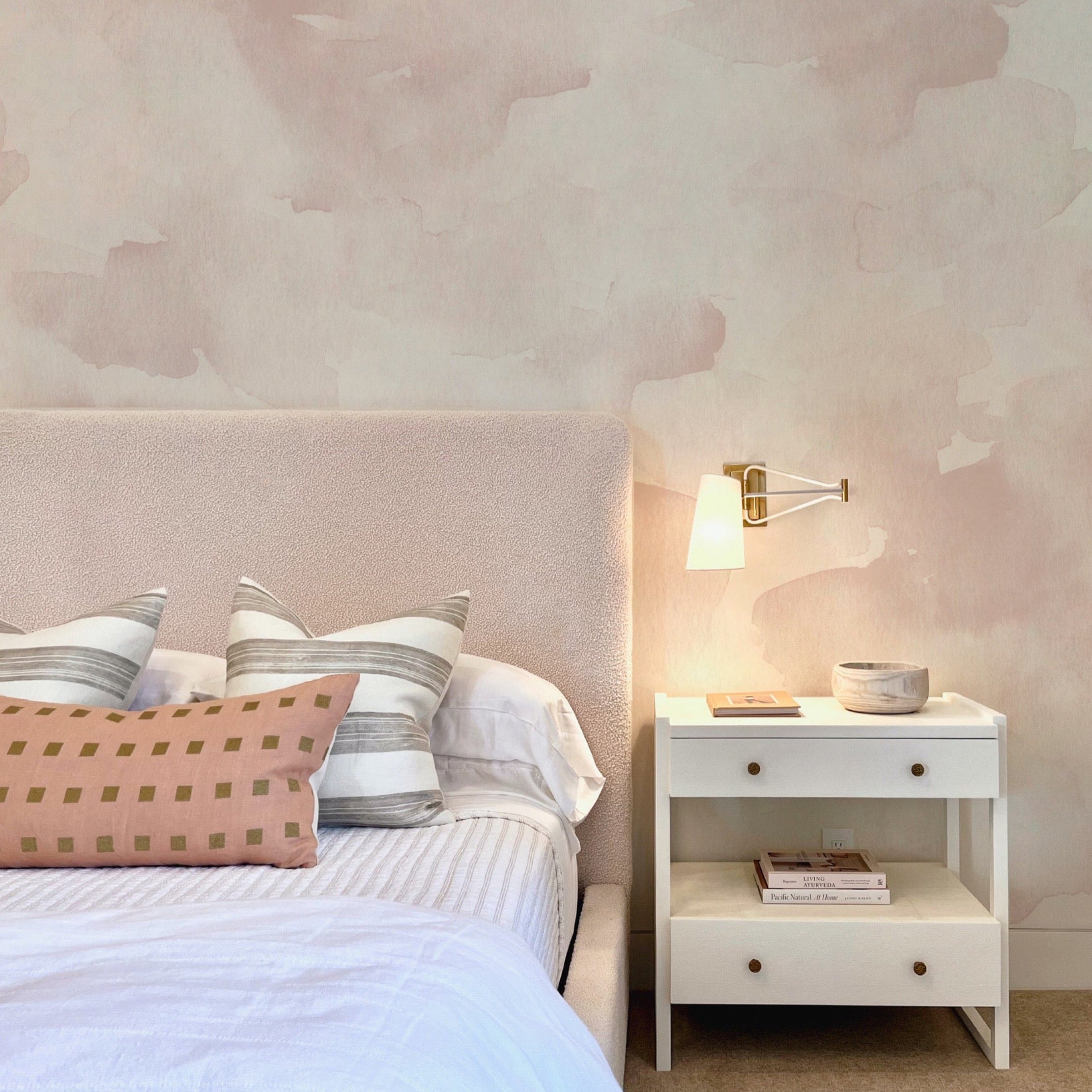 Elegant bedroom interior with Watercolor Mural Wallpaper - Nude Pink, highlighting its soft blush watercolor patterns behind a beige upholstered headboard and a chic bedside table with a modern lamp.