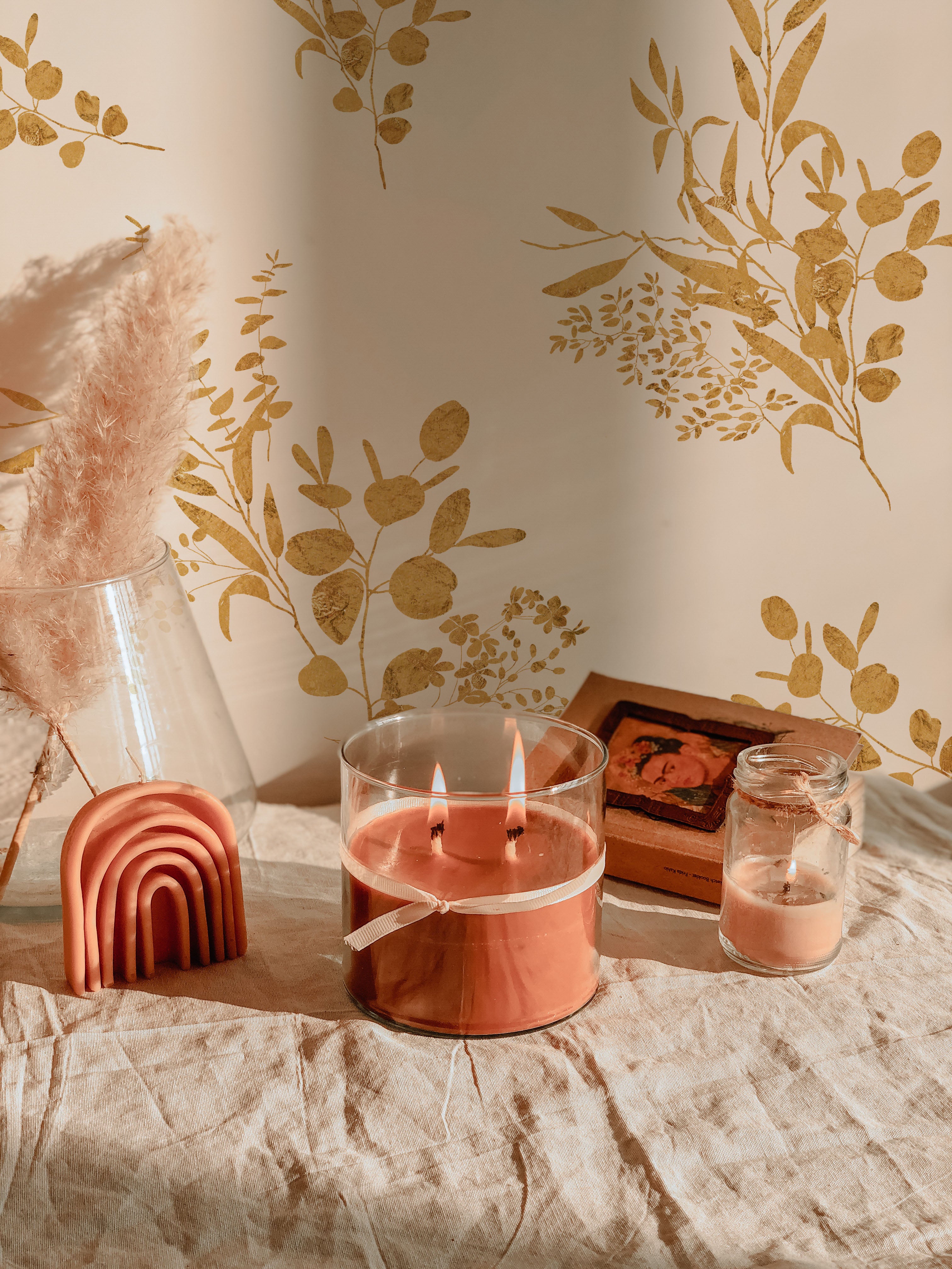 A cozy room setting featuring Golden Greenery Wallpaper with golden botanical patterns on a light beige background, creating a warm and inviting ambiance. The wallpaper accentuates a decor of a large, multi-wick scented candle, a terracotta rainbow-shaped decor item, and vintage books.