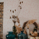 A cozy corner featuring Subtle Botanica Wallpaper III, with its blush-hued leaf patterns adorning the wall behind a vintage wooden dresser topped with vibrant blue glass vases and dried botanicals.